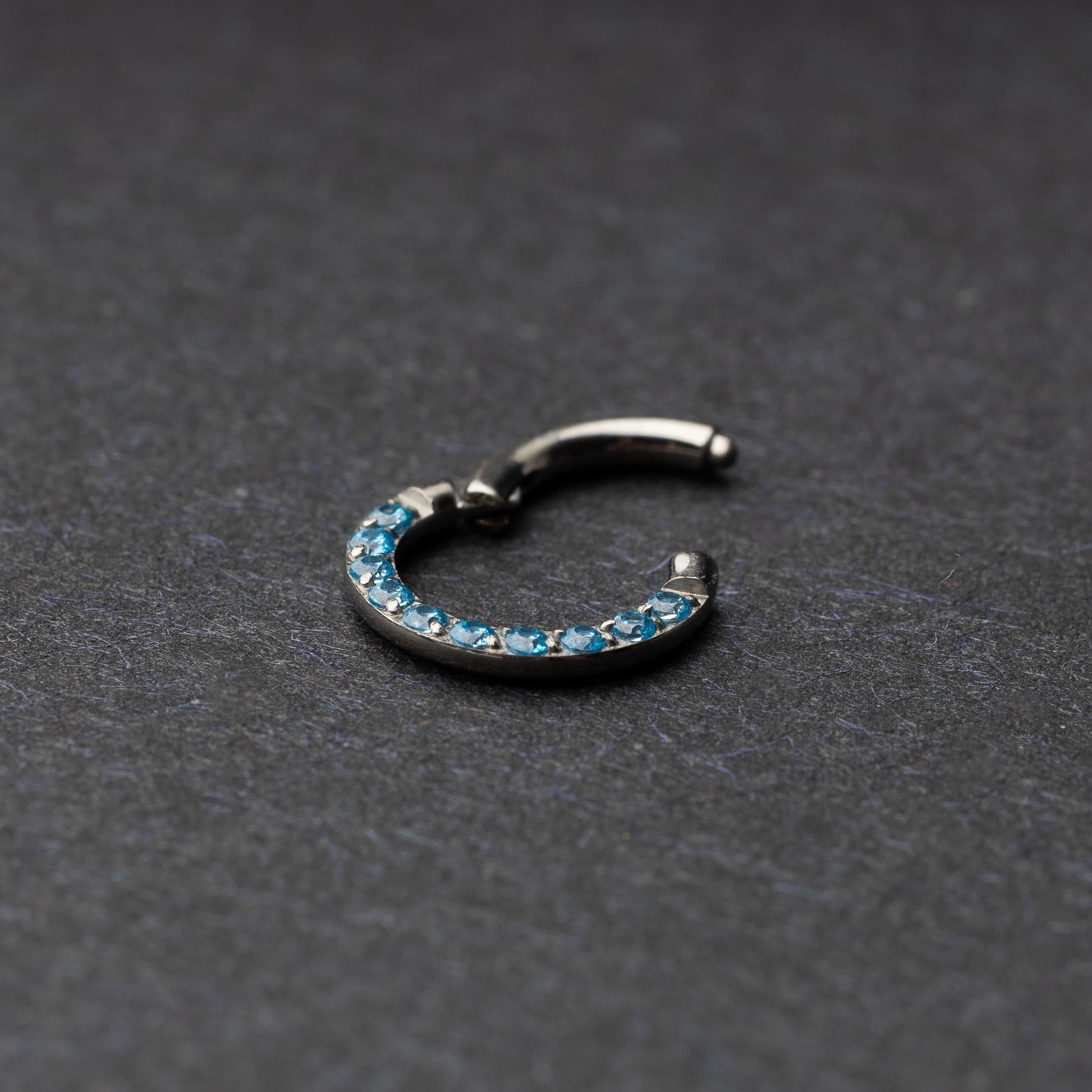 Stainless steel septum clicker with bright blue gemstones, displayed on a dark textured background.