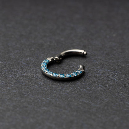 Stainless steel septum clicker with bright blue gemstones, displayed on a dark textured background.