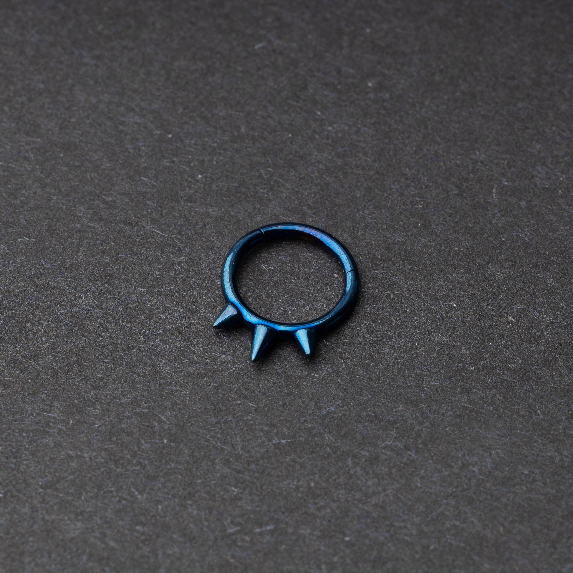 Blue-tone stainless steel clicker ring with three spikes, displayed on a dark textured background.