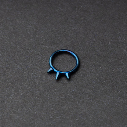 Blue-tone stainless steel clicker ring with three spikes, displayed on a dark textured background.