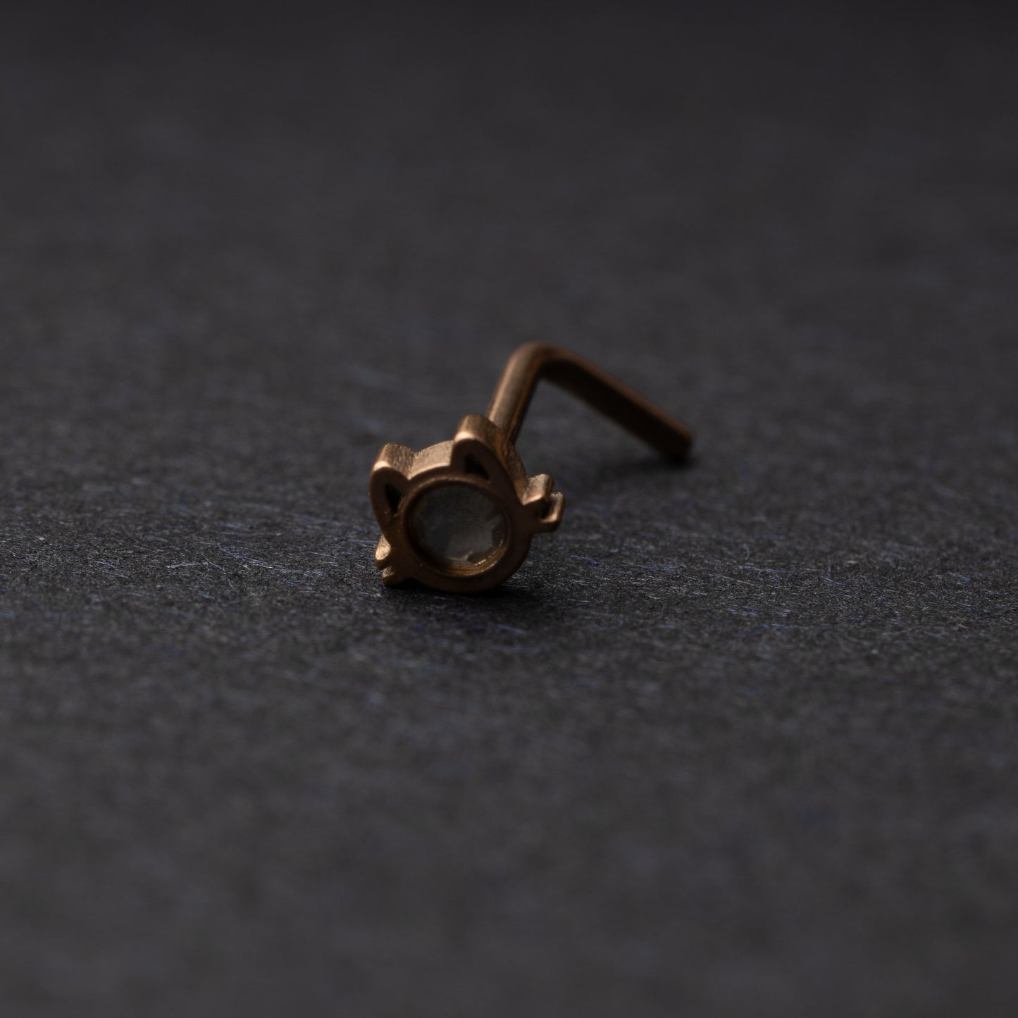 A rose gold stud earring in the shape of a cat’s face with a bezel-set gemstone, displayed on a dark background.