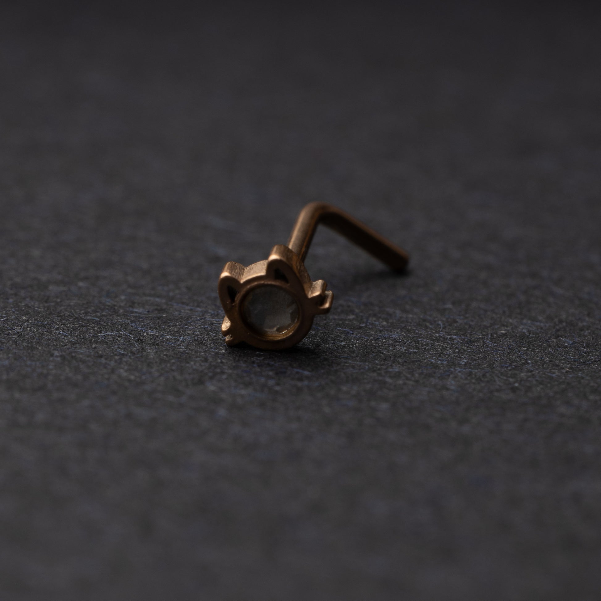 A rose gold stud earring in the shape of a cat’s face with a bezel-set gemstone, displayed on a dark background.
