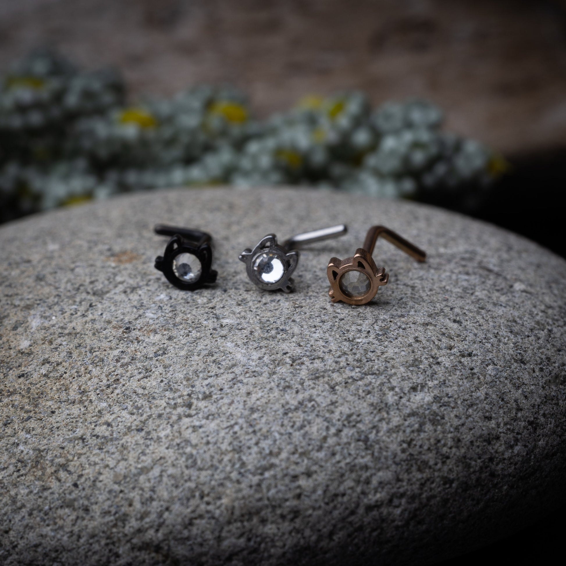 Black, silver and rose gold stud earrings in the shape of a cat’s face with a bezel-set gemstone, displayed on a stone background