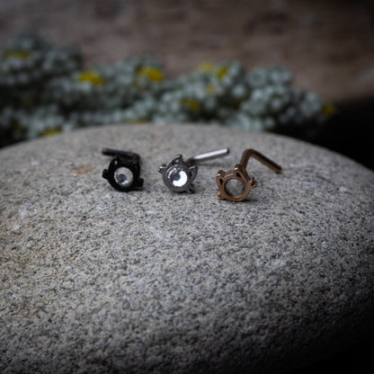 Black, silver and rose gold stud earrings in the shape of a cat’s face with a bezel-set gemstone, displayed on a stone background