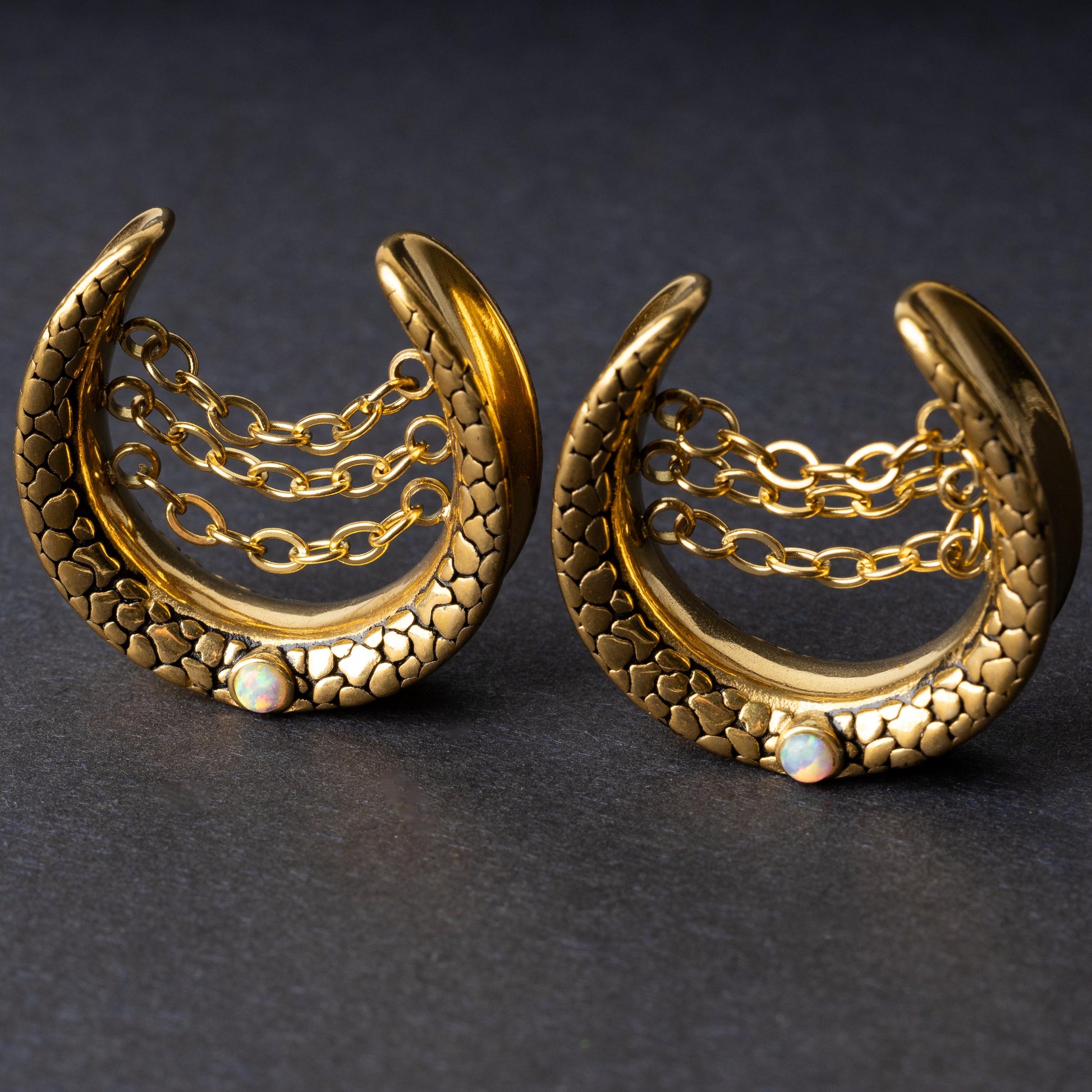 Pair of crescent-shaped hoops in gold with textured details, cascading chains, and an opal accent at the base, displayed on a dark background.