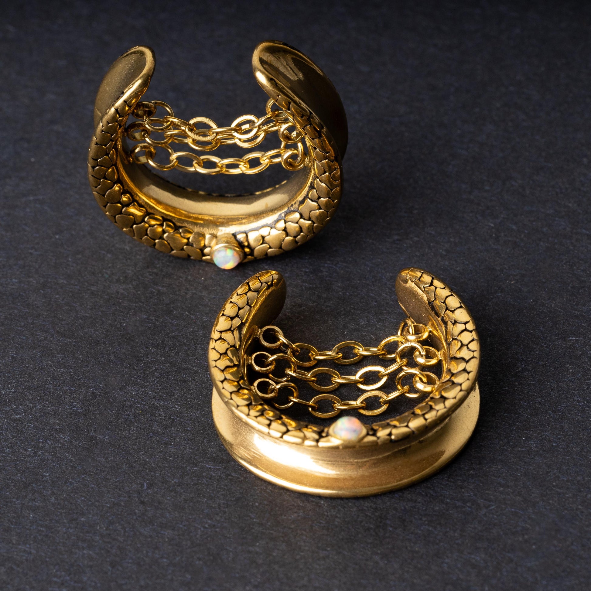 A pair of crescent-shaped hoops in gold with textured details, cascading chains, and an opal accent at the base, displayed on a dark background.