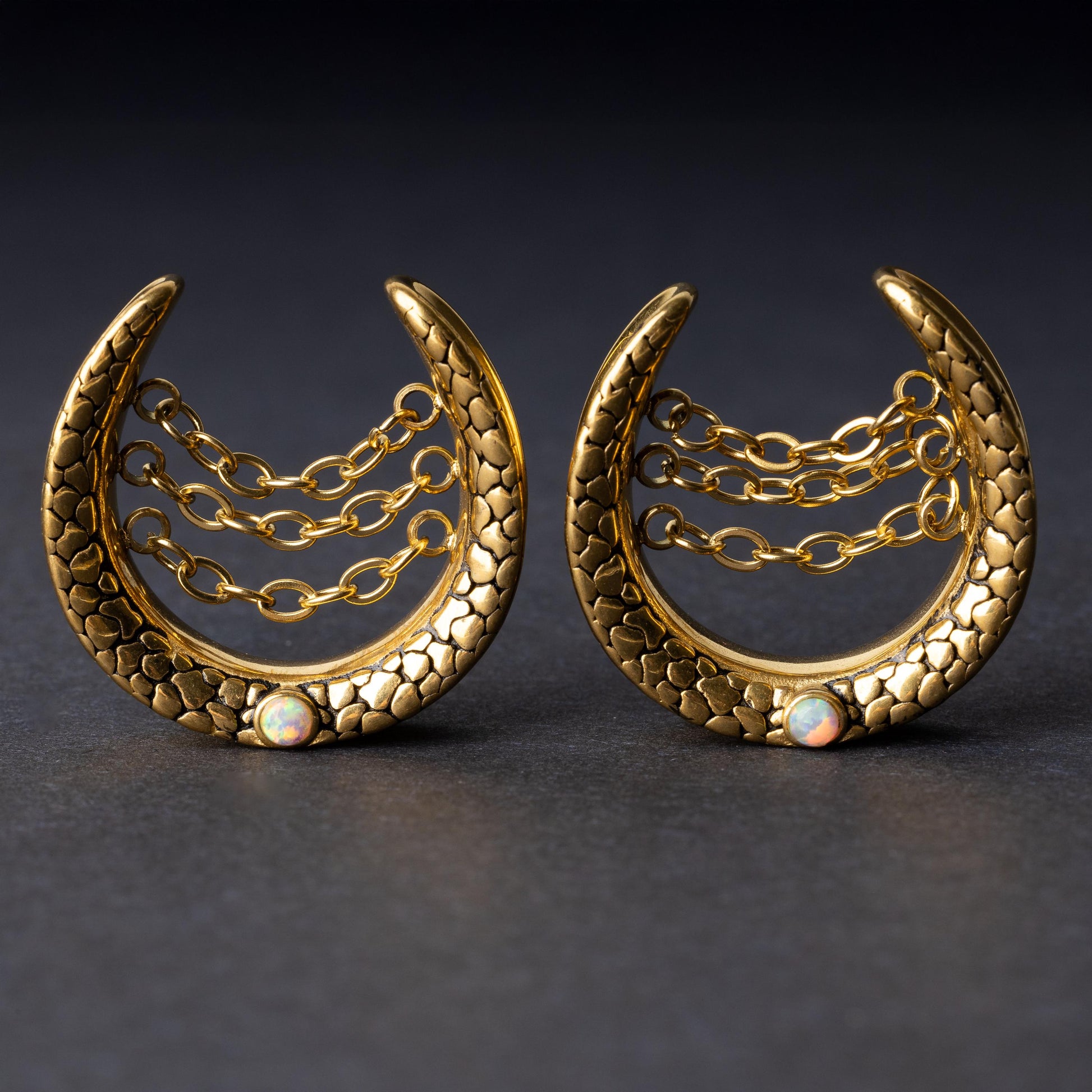 Pair of crescent-shaped hoops in gold with textured details, cascading chains, and an opal accent at the base, displayed on a dark background.