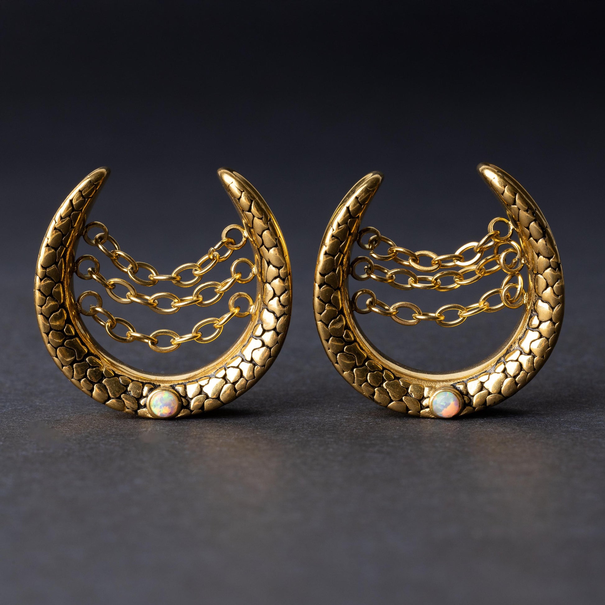 Pair of crescent-shaped hoops in gold with textured details, cascading chains, and an opal accent at the base, displayed on a dark background.
