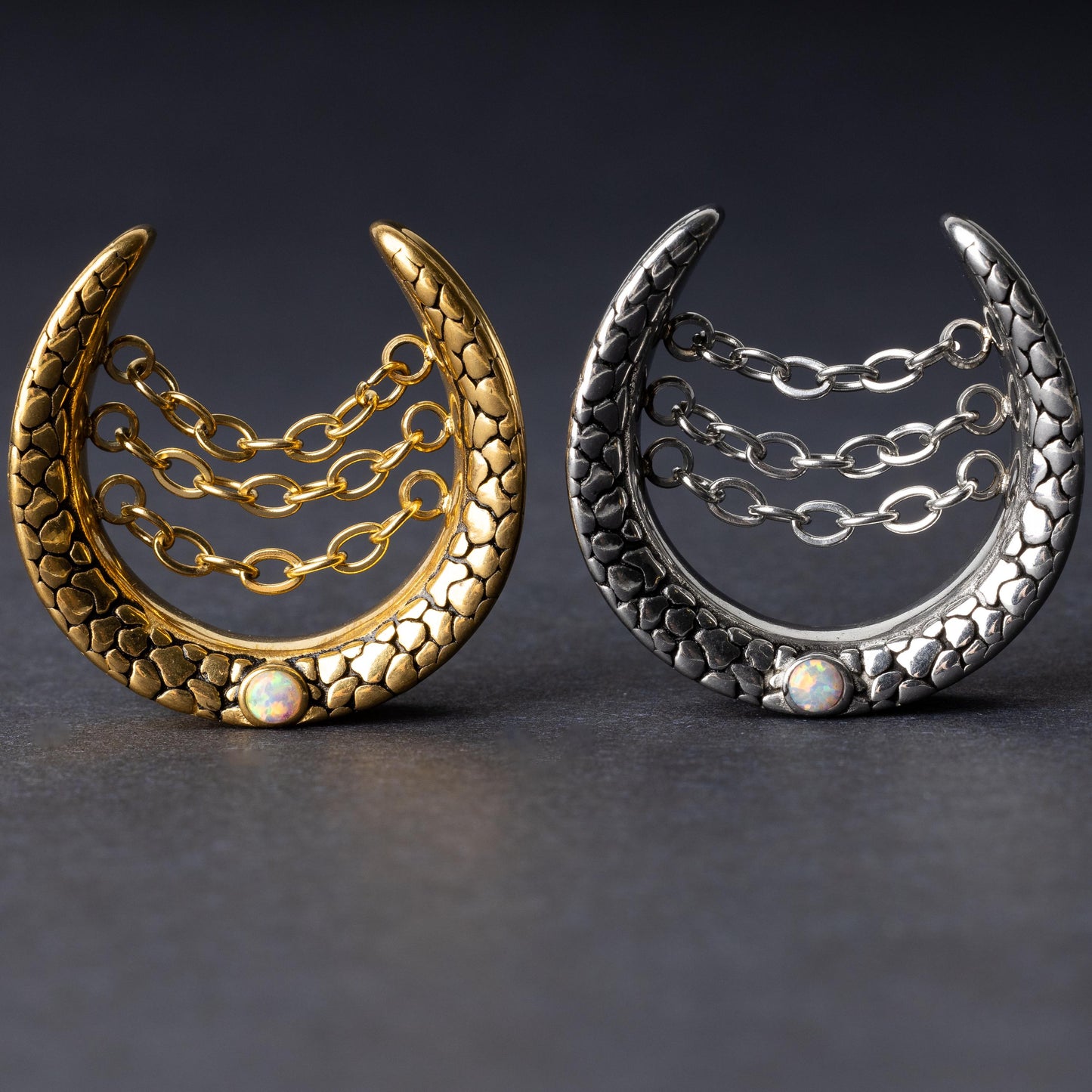 Crescent-shaped hoops in gold and silver finishes with textured details, cascading chains, and an opal accent at the base, displayed on a dark background.