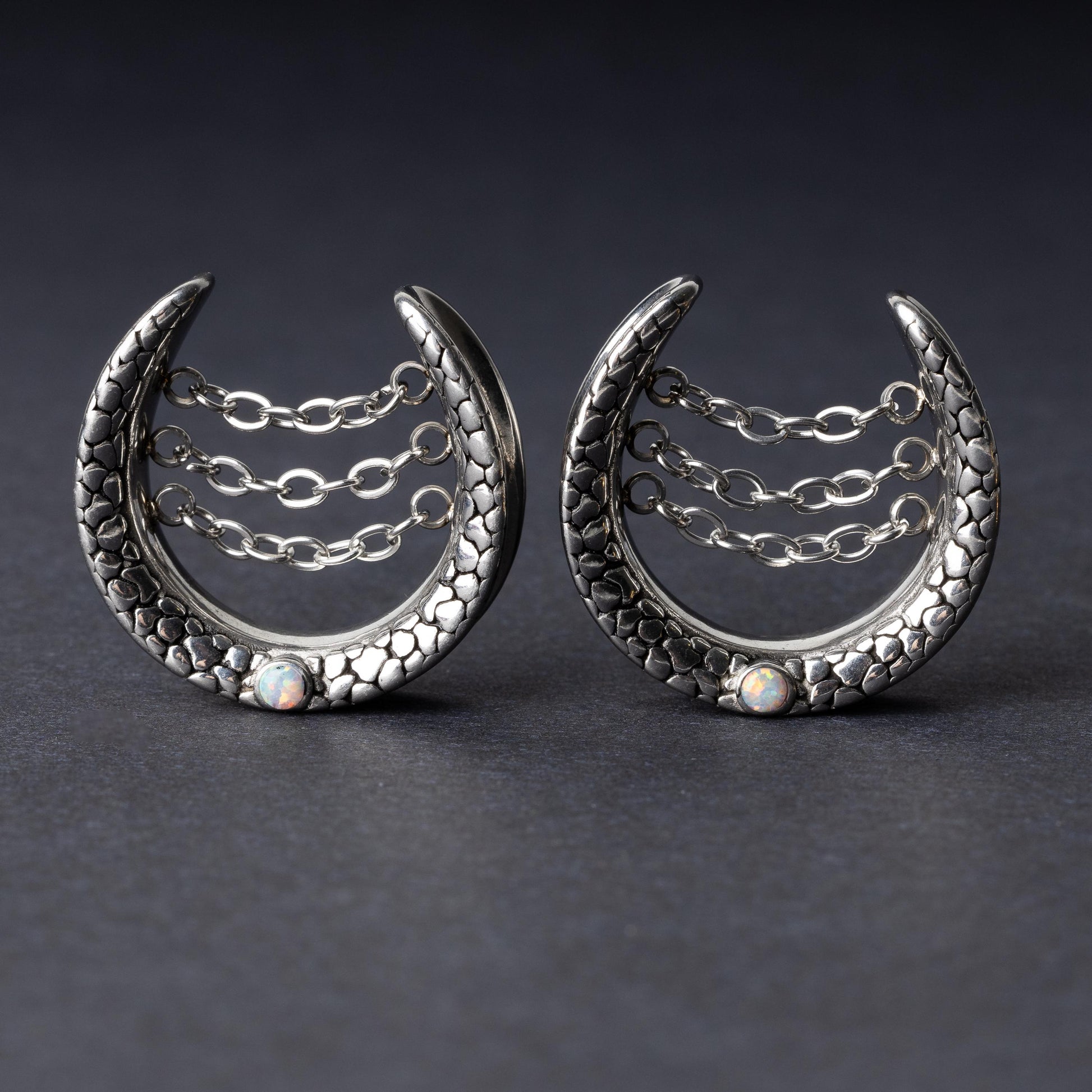 Pair of crescent-shaped hoops in silver finish with textured details, cascading chains, and an opal accent at the base, displayed on a dark background.