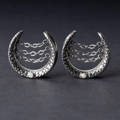 Pair of crescent-shaped hoops in silver finish with textured details, cascading chains, and an opal accent at the base, displayed on a dark background.