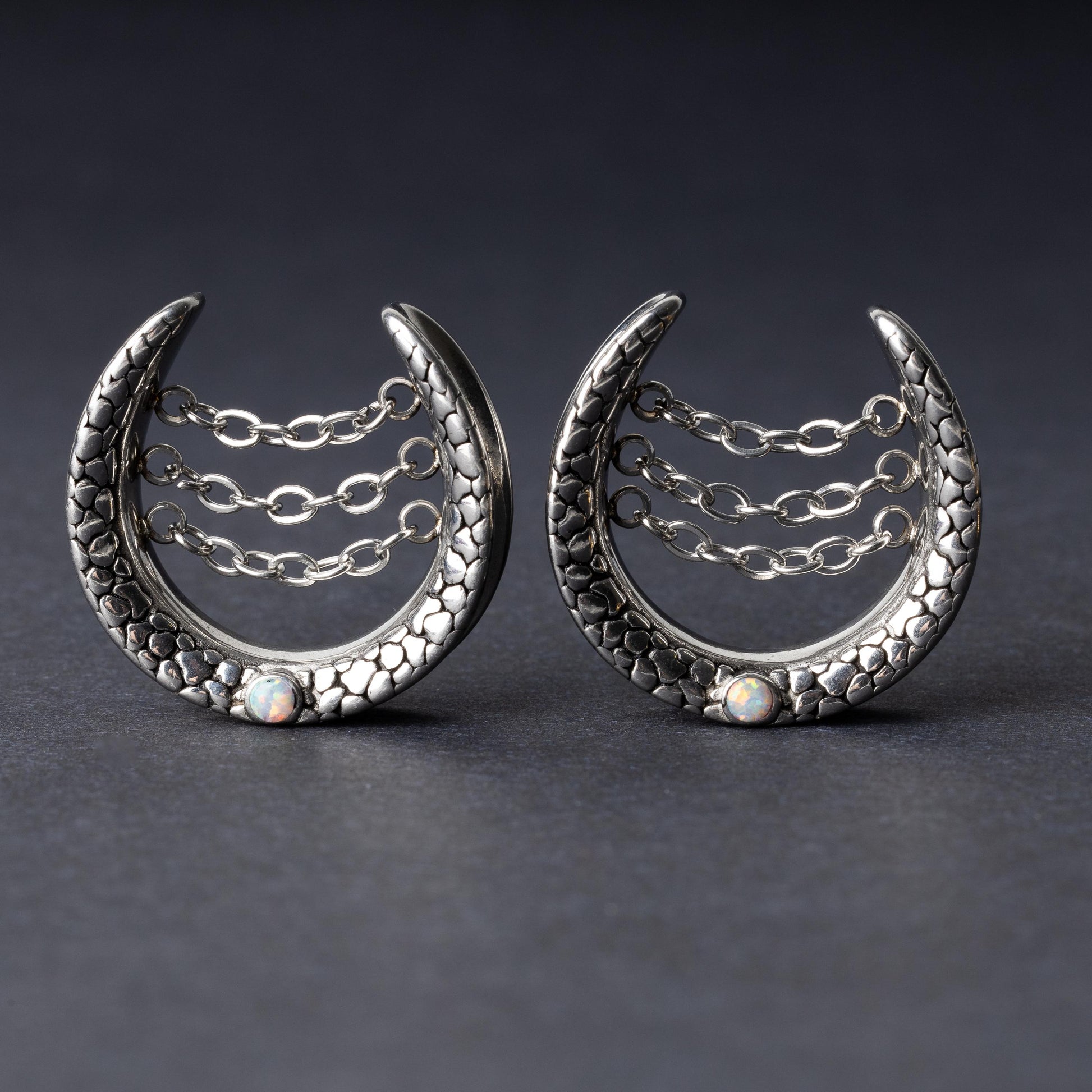 Pair of crescent-shaped hoops in silver with textured details, cascading chains, and an opal accent at the base, displayed on a dark background.