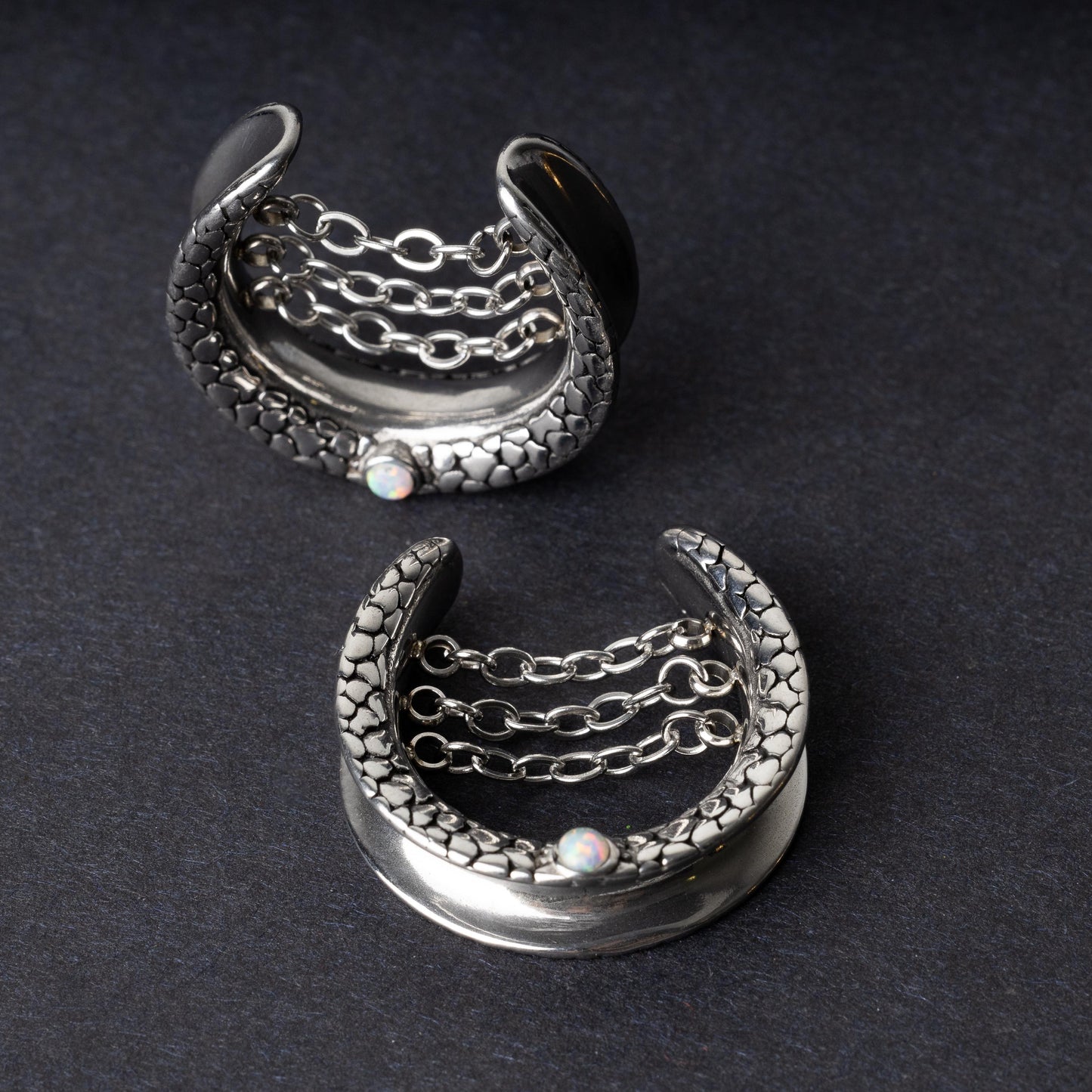 A pair of crescent-shaped hoops silver with textured details, cascading chains, and an opal accent at the base, displayed on a dark background.