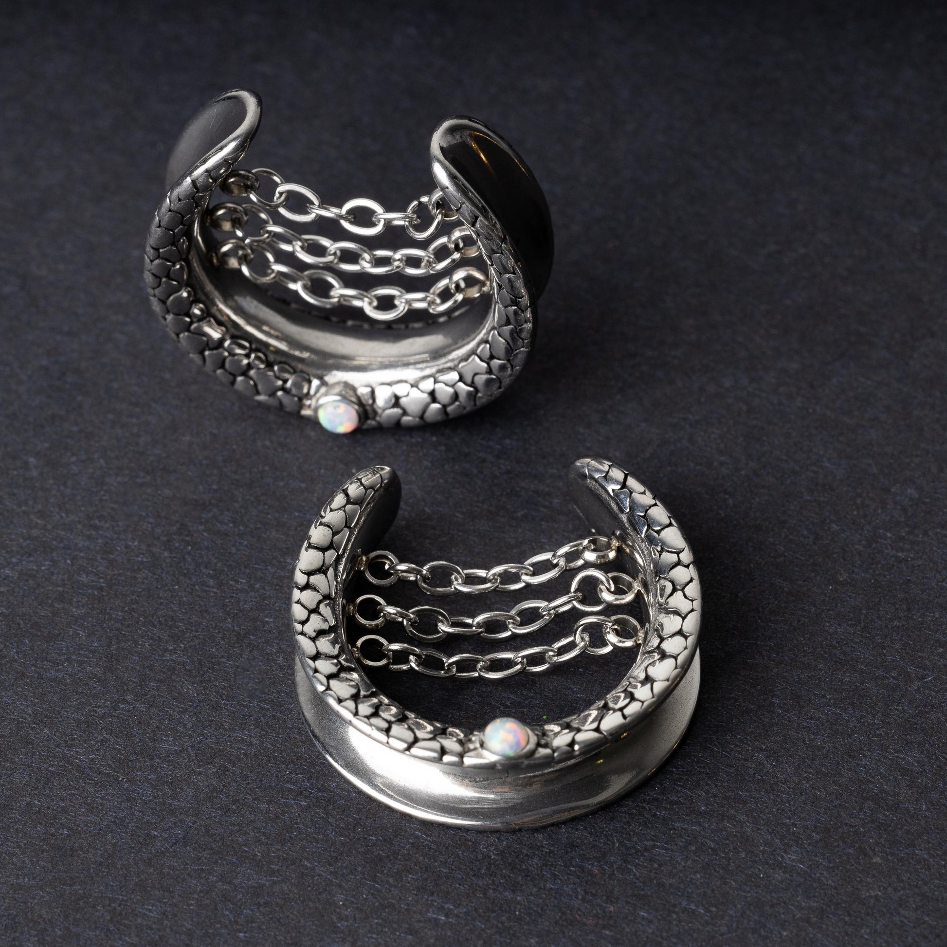 A pair of crescent-shaped hoops silver with textured details, cascading chains, and an opal accent at the base, displayed on a dark background.
