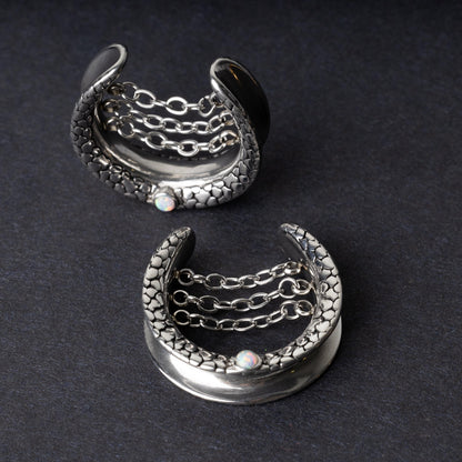 A pair of crescent-shaped hoops silver with textured details, cascading chains, and an opal accent at the base, displayed on a dark background.