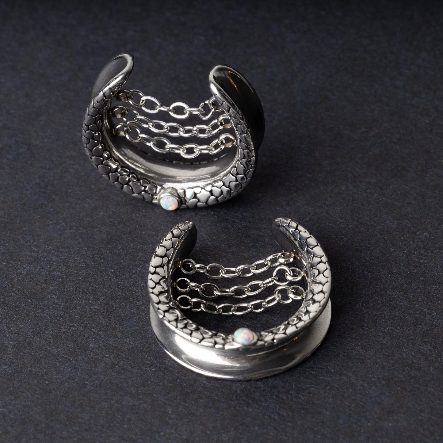 A pair of crescent-shaped hoops silver with textured details, cascading chains, and an opal accent at the base, displayed on a dark background.