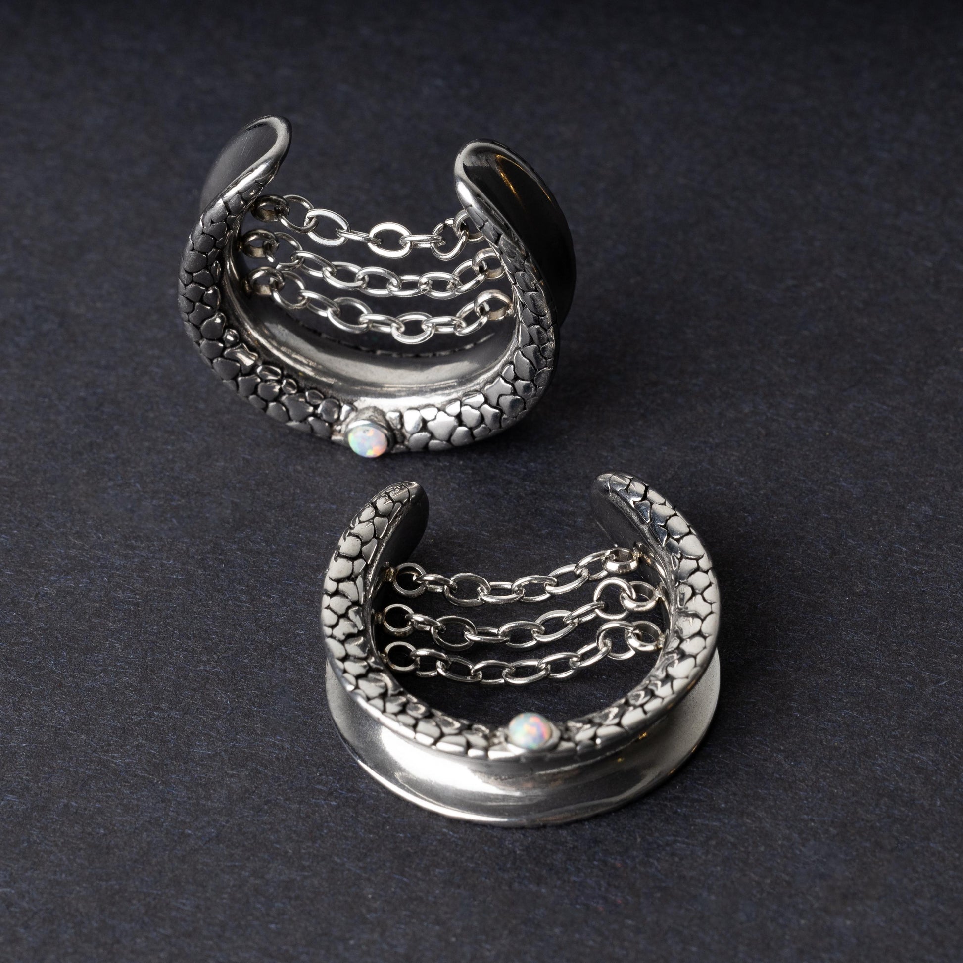 A pair of crescent-shaped hoops silver with textured details, cascading chains, and an opal accent at the base, displayed on a dark background.