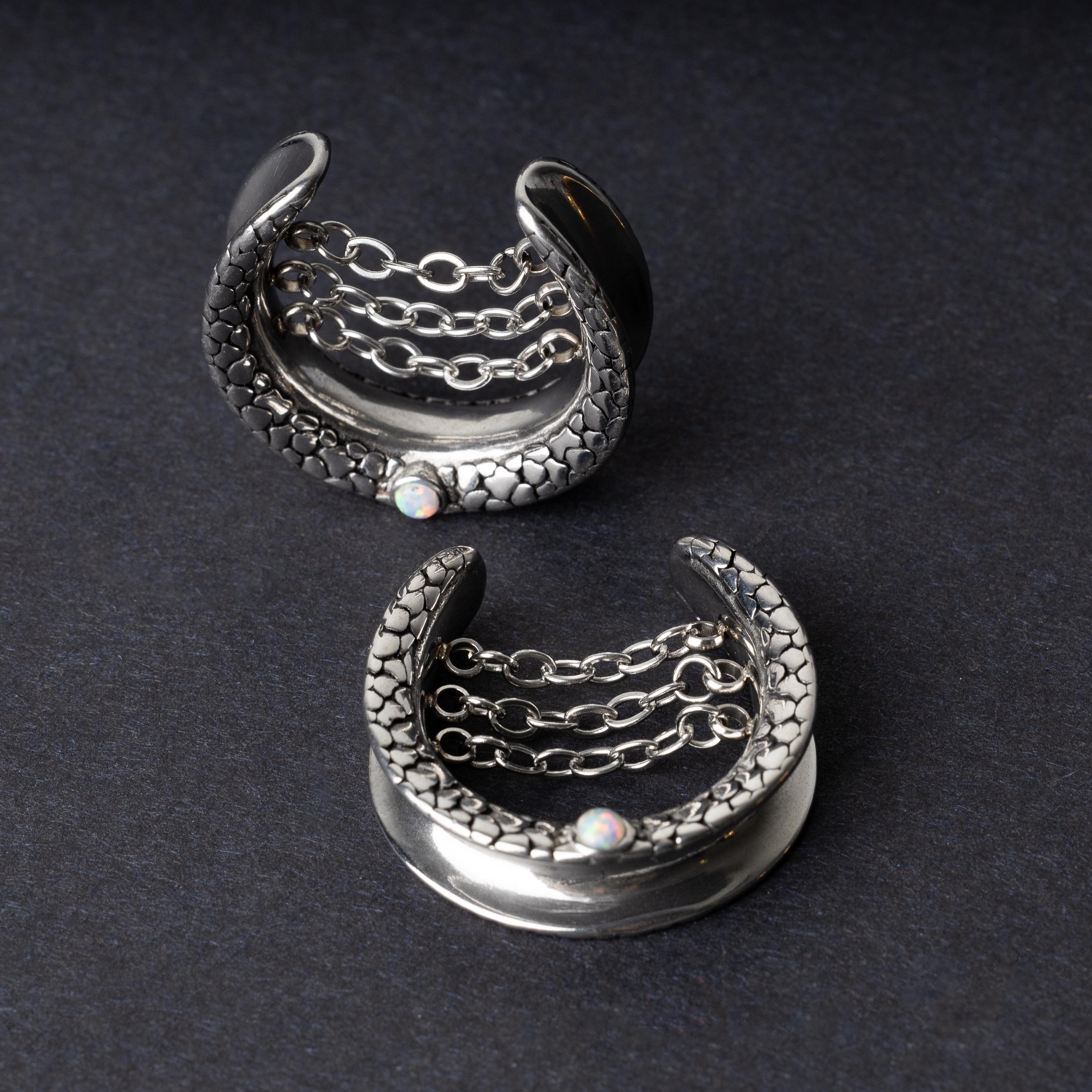 A pair of crescent-shaped hoops silver with textured details, cascading chains, and an opal accent at the base, displayed on a dark background.