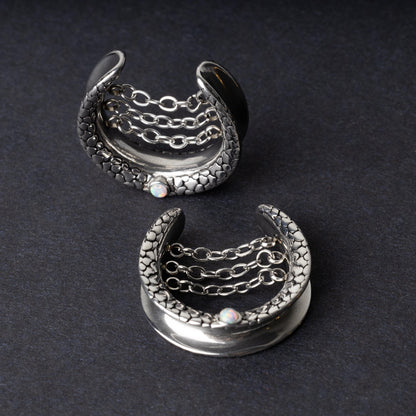 A pair of crescent-shaped hoops silver with textured details, cascading chains, and an opal accent at the base, displayed on a dark background.