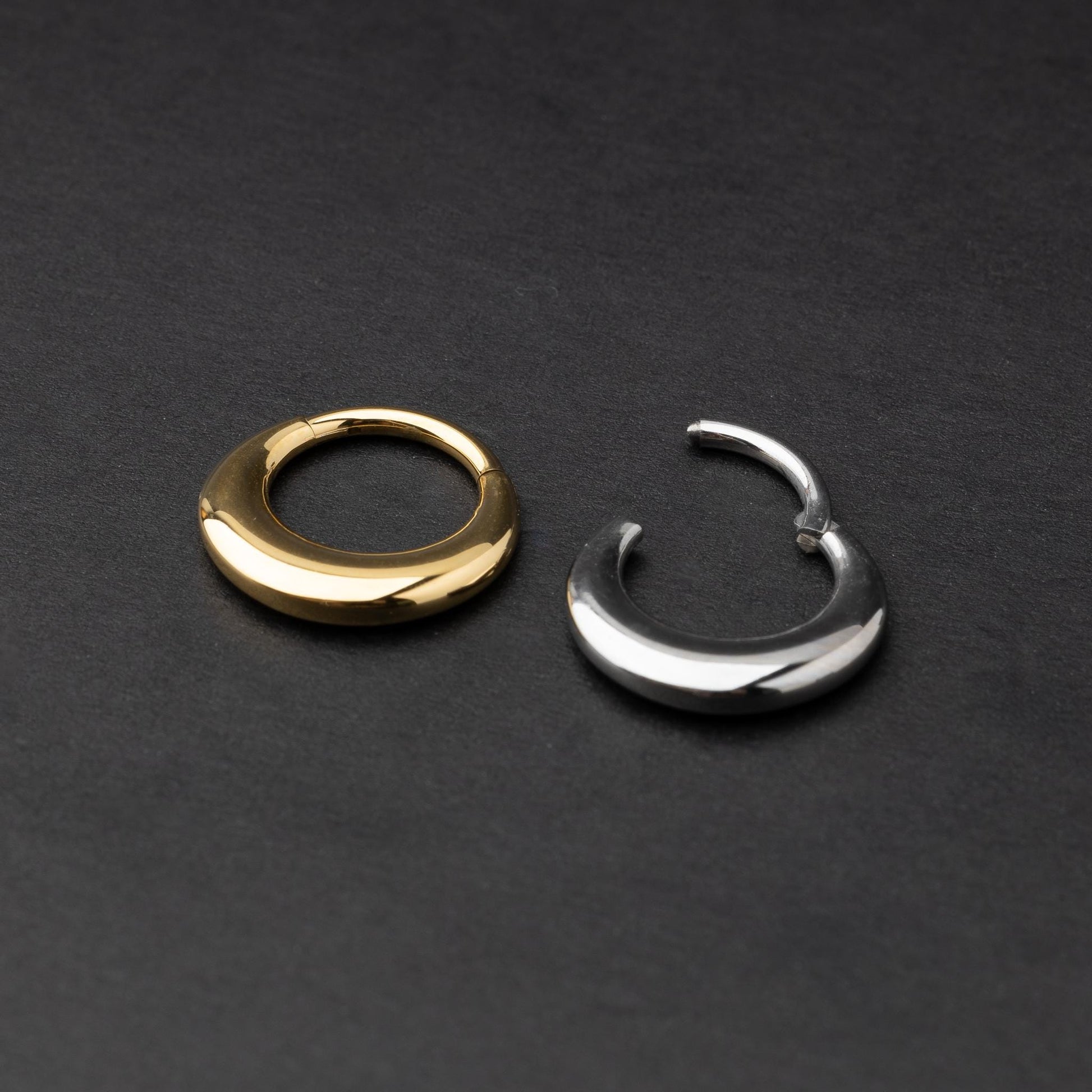 Crescent-shaped septum clickers, one in gold and one in silver, with a polished finish, displayed on a dark background.
