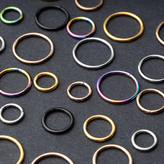 A variety of individually sold seamless clicker hoops in different sizes and finishes, including gold, rose gold, silver, black, and rainbow, displayed on a dark background.