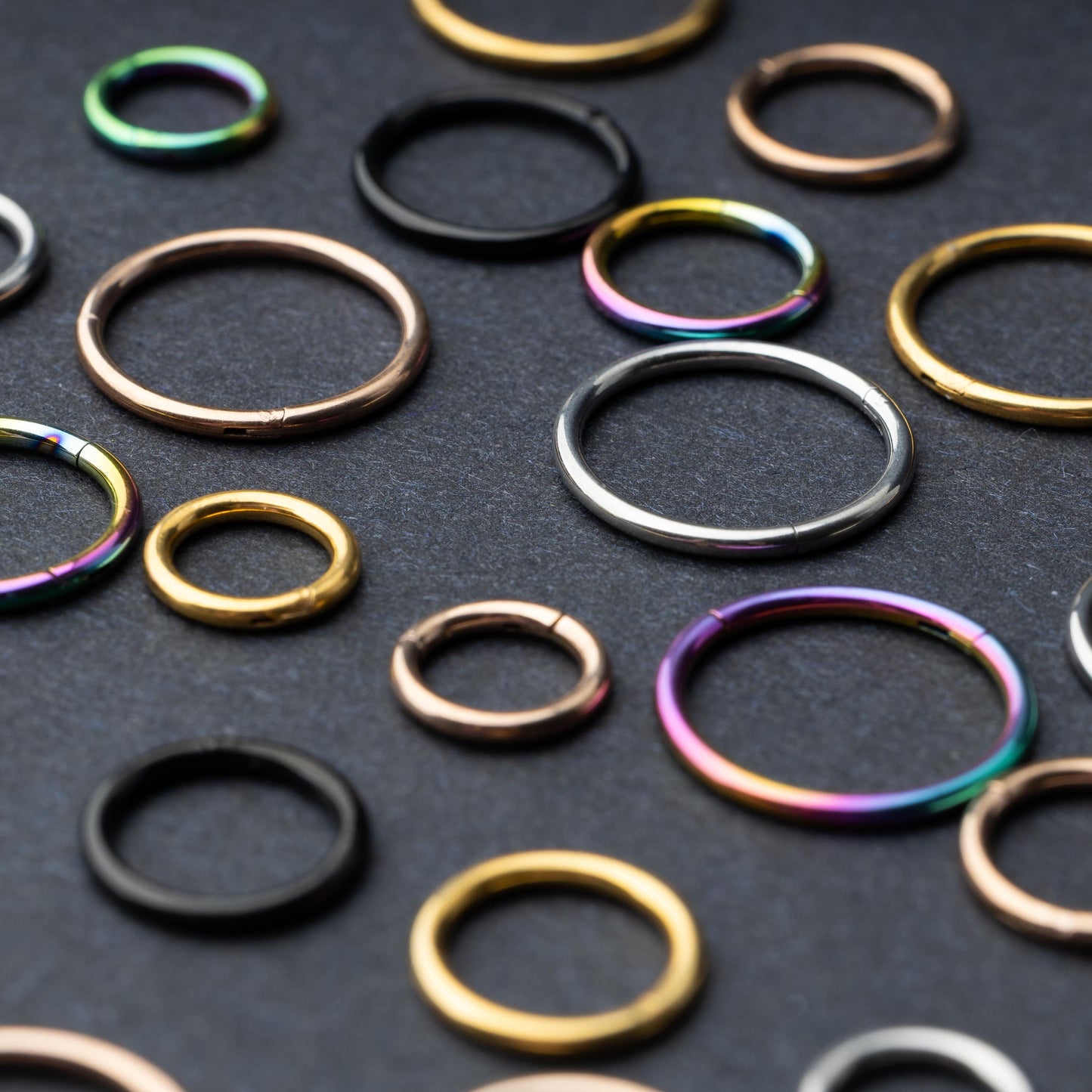 A variety of individually sold seamless clicker hoops in different sizes and finishes, including gold, rose gold, silver, black, and rainbow, displayed on a dark background.