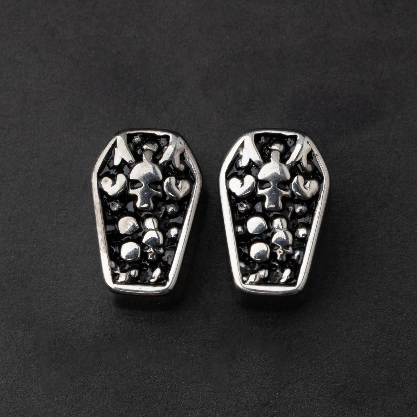 A pair of coffin-shaped stainless steel ear plugs with intricate skeleton designs, displayed on a dark background.