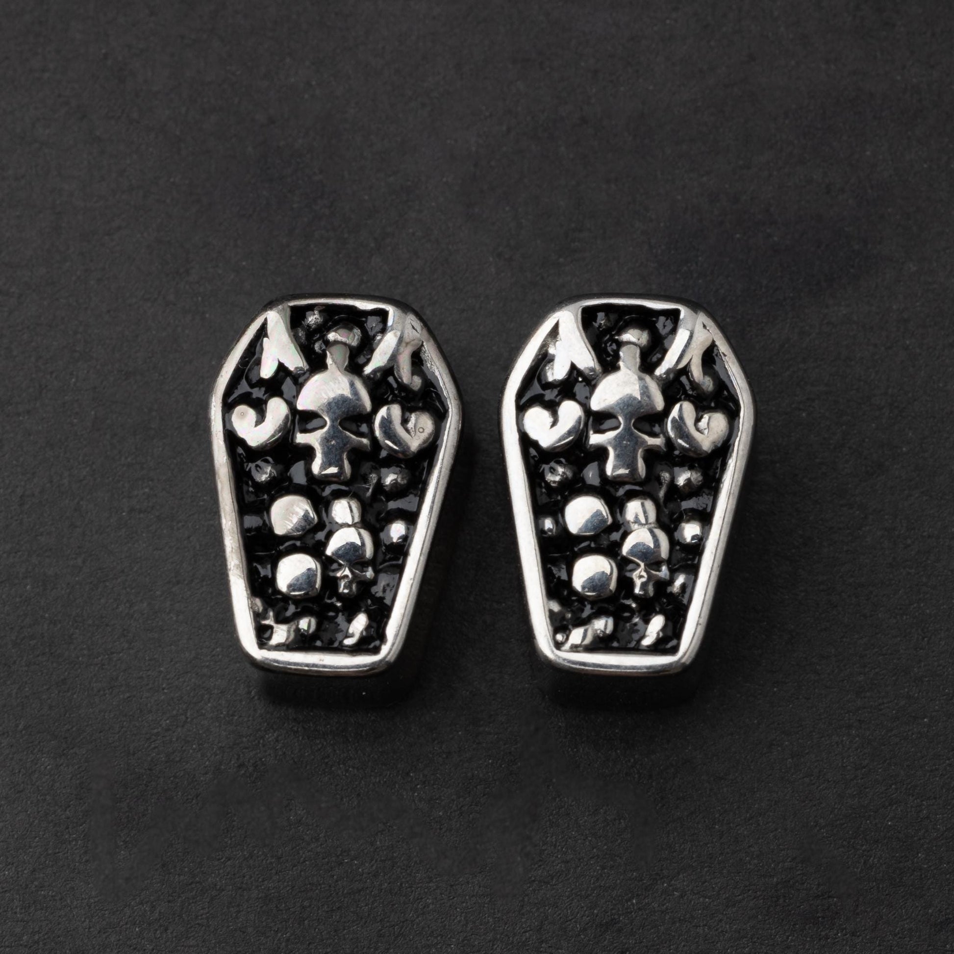 A pair of coffin-shaped stainless steel ear plugs with intricate skeleton designs, displayed on a dark background.
