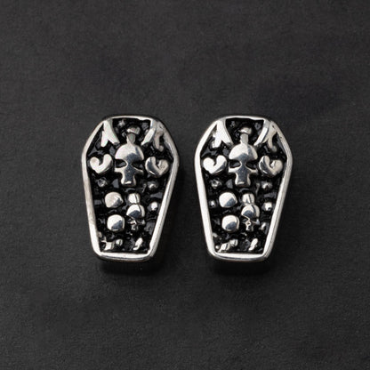 A pair of coffin-shaped stainless steel ear plugs with intricate skeleton designs, displayed on a dark background.