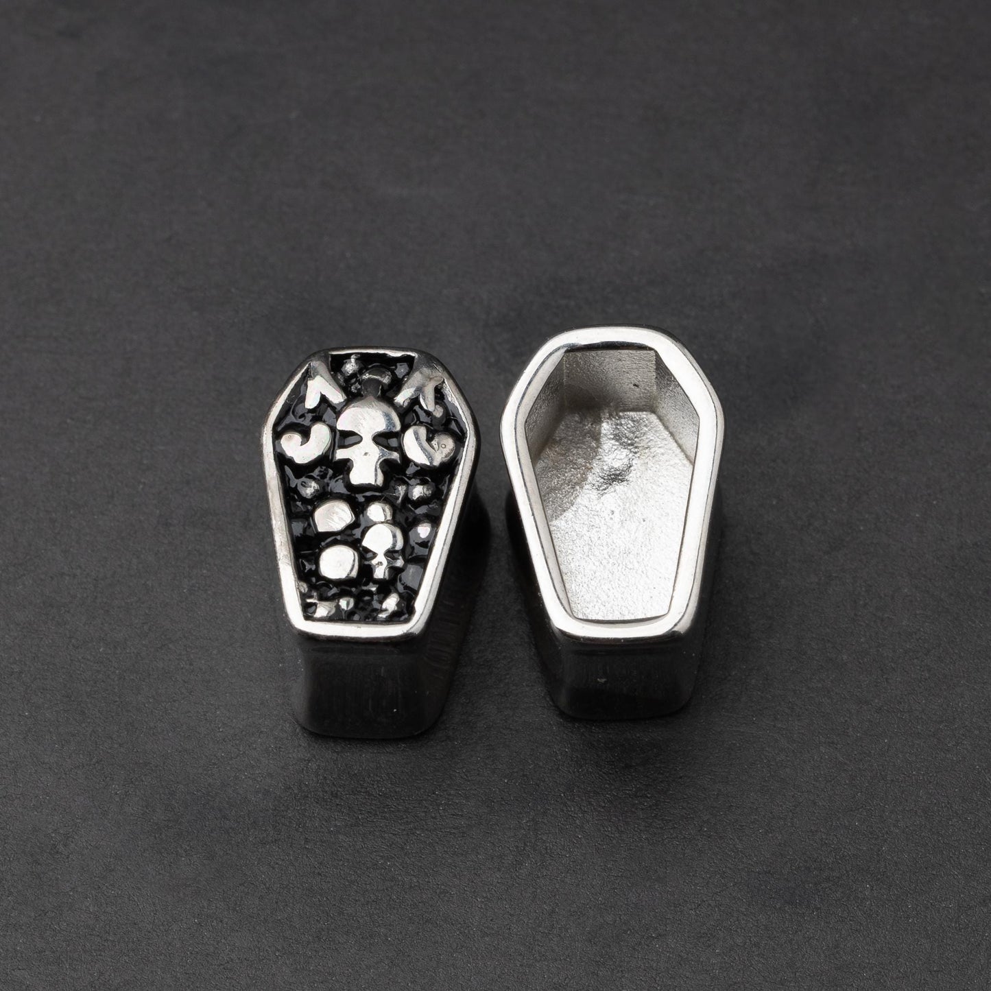 A pair of coffin-shaped stainless steel ear plugs with intricate skeleton designs, displayed on a dark background.