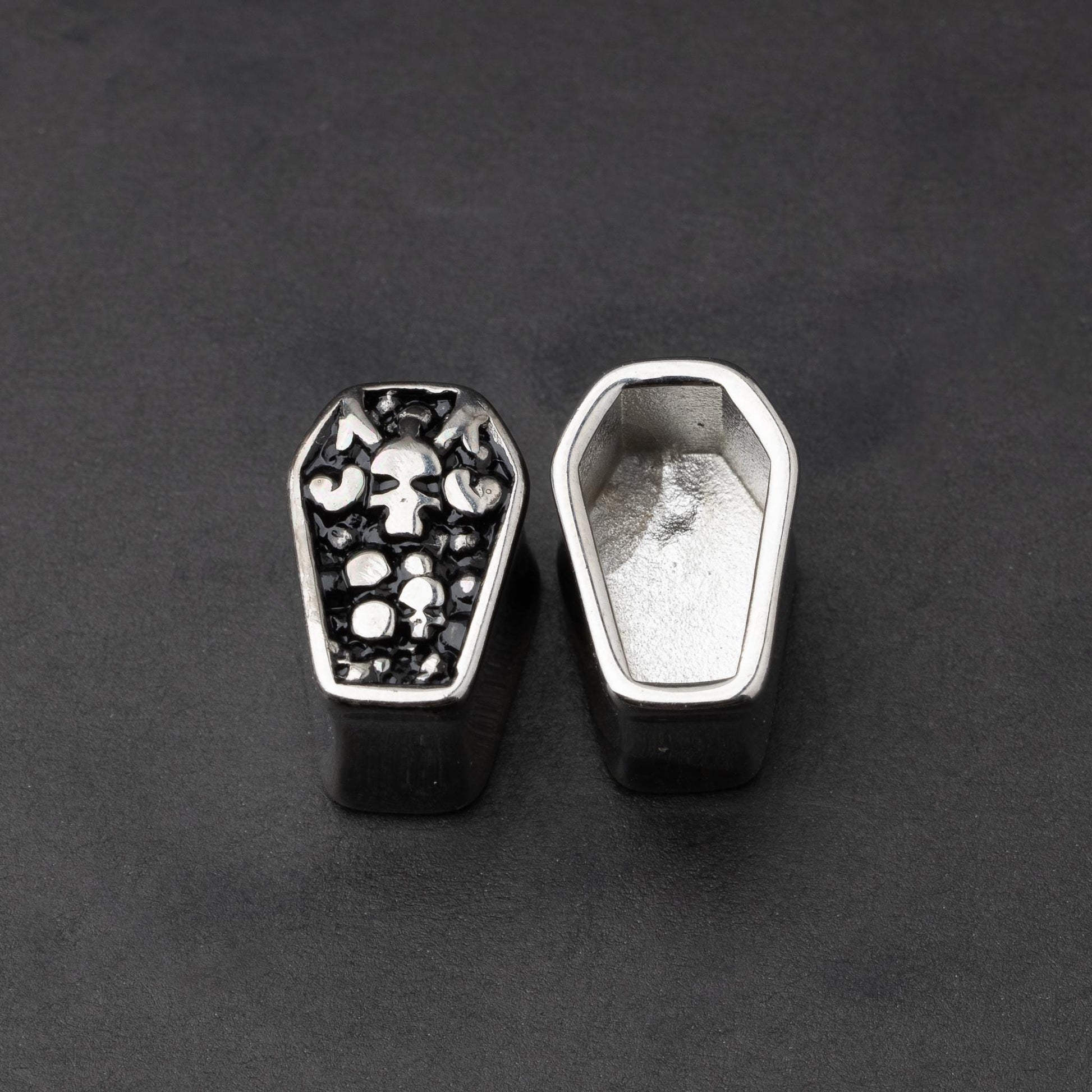 A pair of coffin-shaped stainless steel ear plugs with intricate skeleton designs, displayed on a dark background.