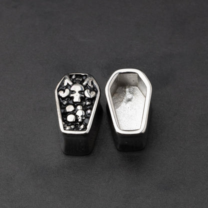 A pair of coffin-shaped stainless steel ear plugs with intricate skeleton designs, displayed on a dark background.