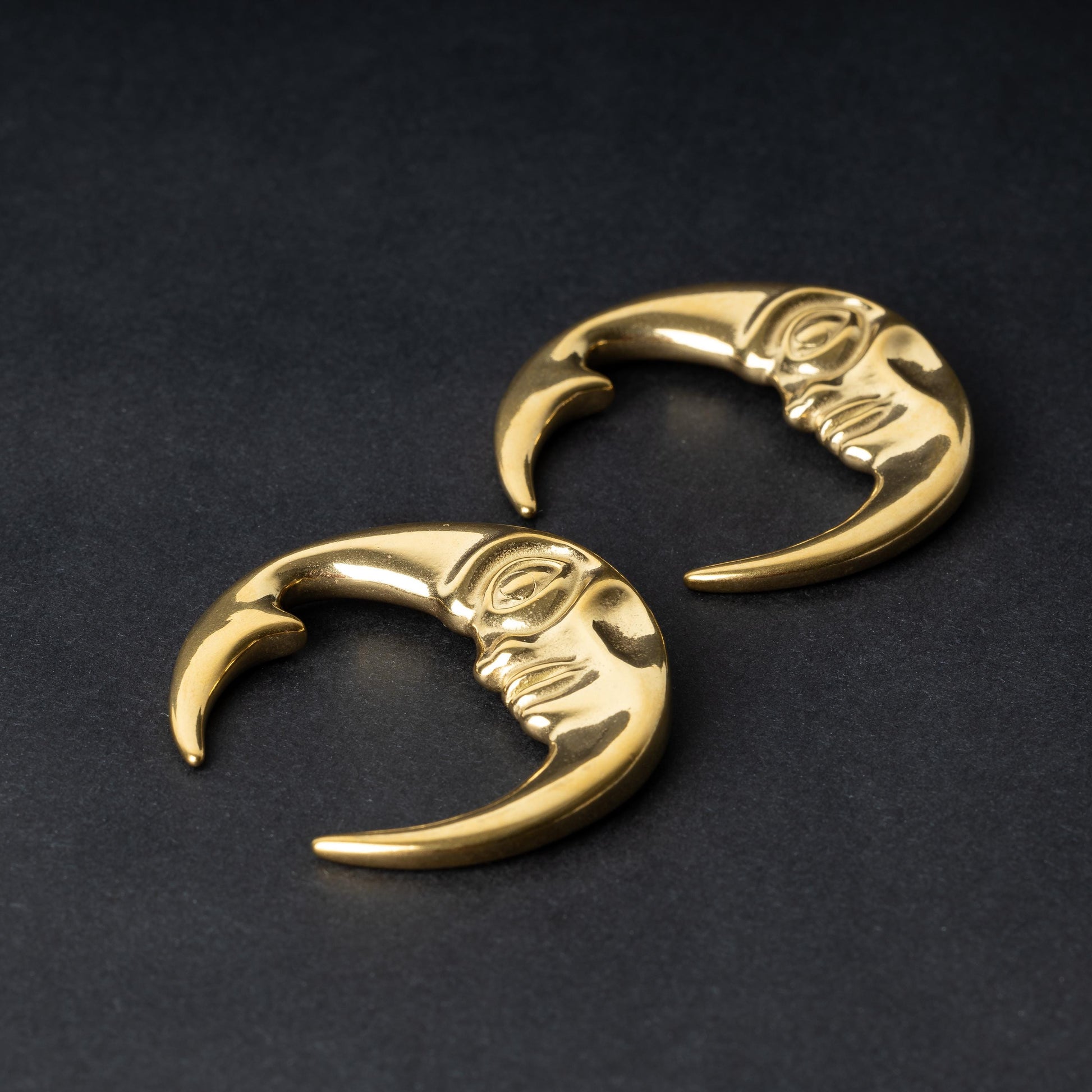 A pair of crescent moon-shaped ear weights in polished gold, featuring a detailed moon face design, displayed on a dark background.

