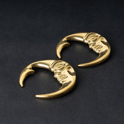 A pair of crescent moon-shaped ear weights in polished gold, featuring a detailed moon face design, displayed on a dark background.

