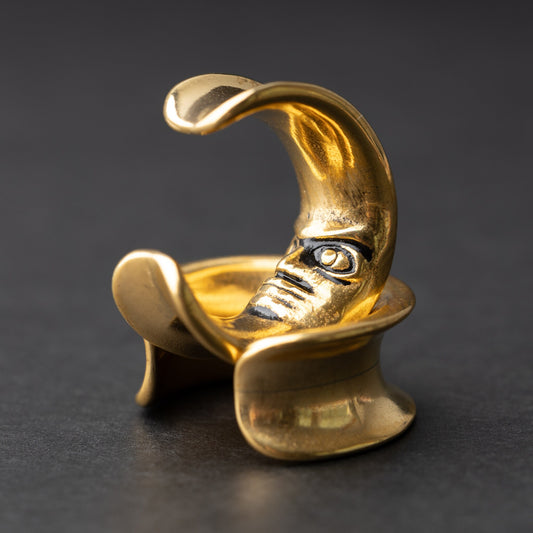 Close-up of a gold crescent moon-shaped ear weight with a detailed face design, displayed on a dark background.