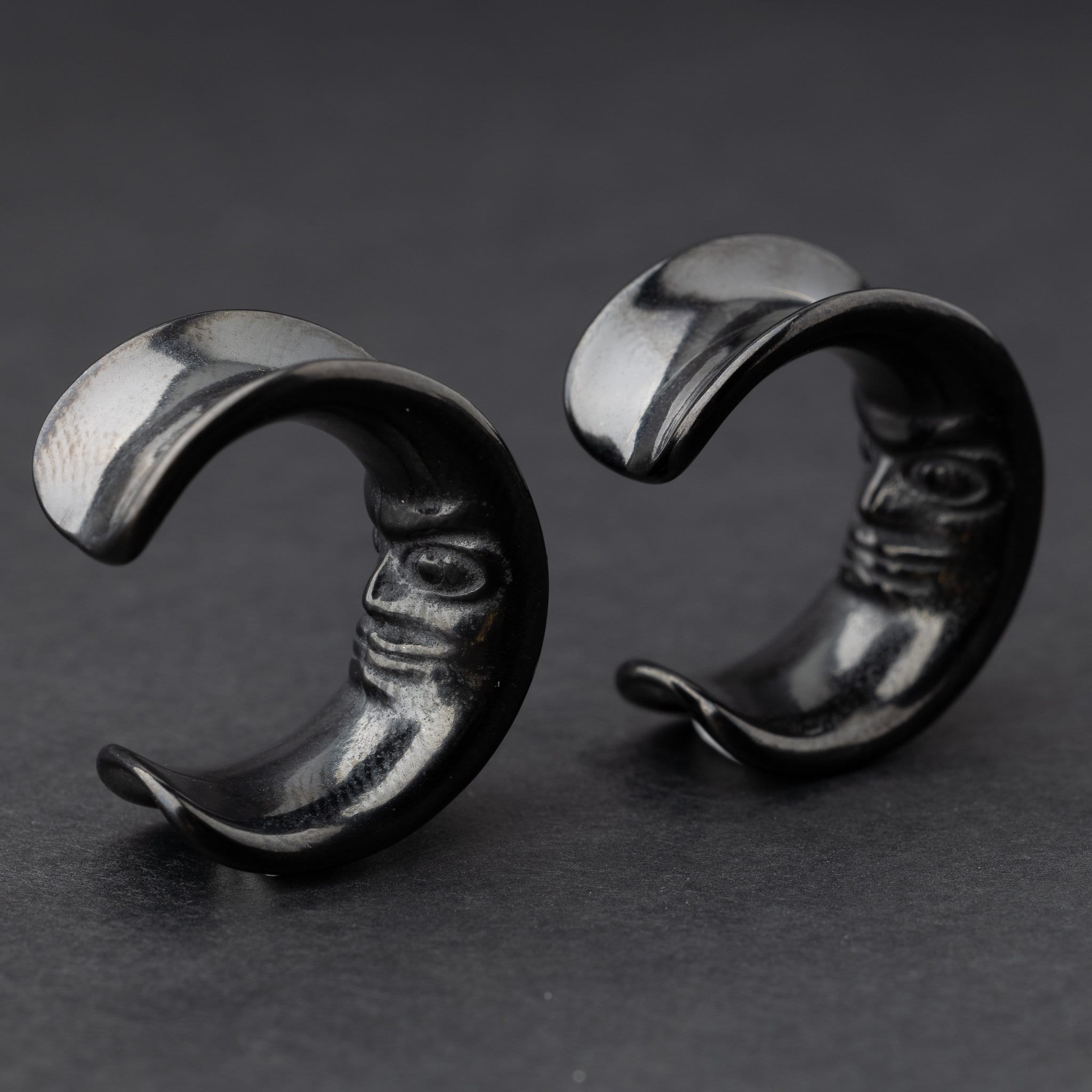 Close-up of a pair of black crescent moon-shaped ear weights with a detailed face design, displayed on a dark background.