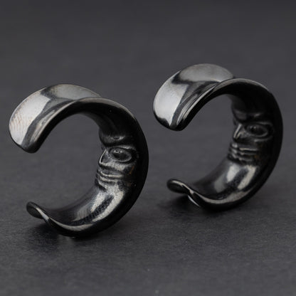 Close-up of a pair of black crescent moon-shaped ear weights with a detailed face design, displayed on a dark background.