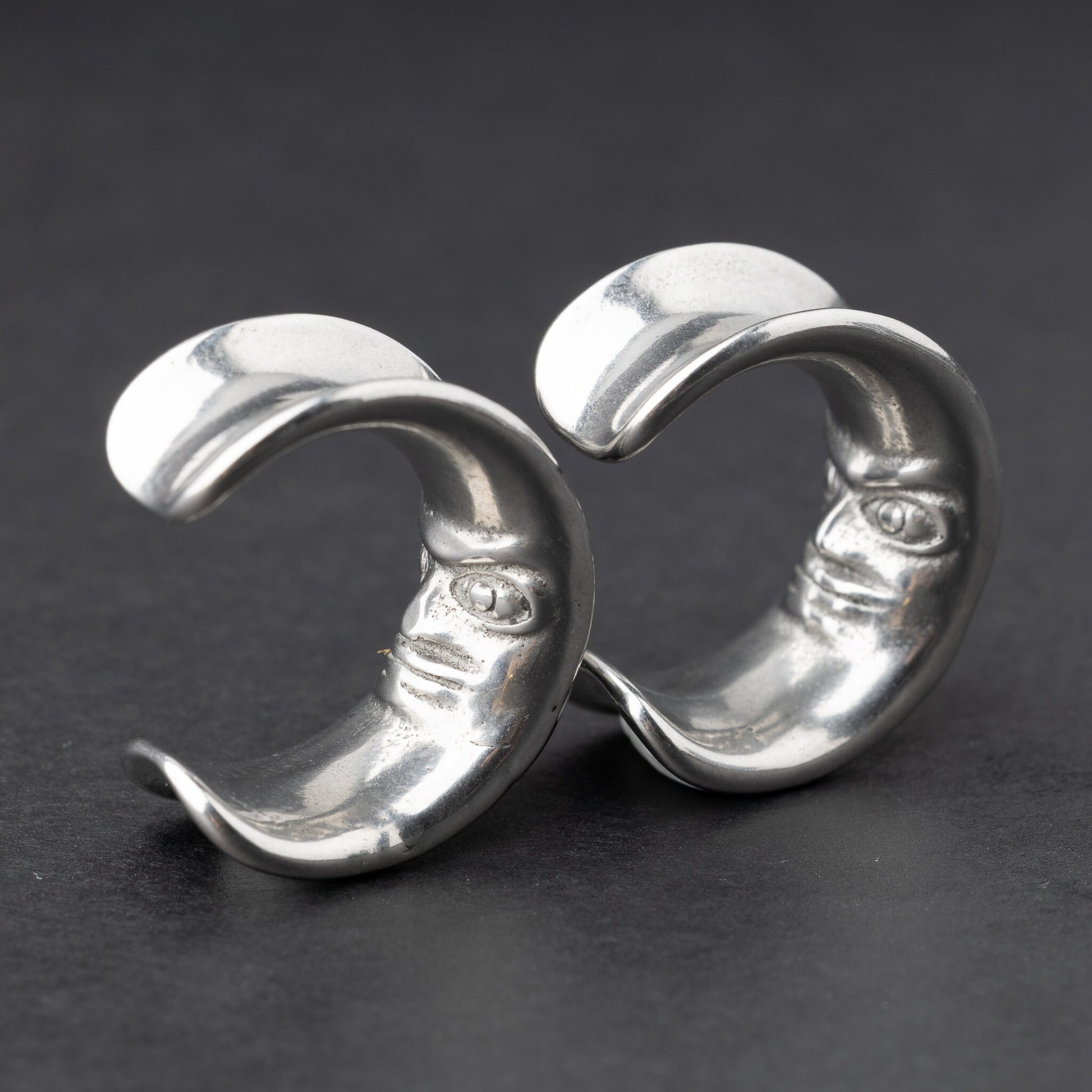 Close-up of a pair of silver crescent moon-shaped ear weights with a detailed face design, displayed on a dark background.