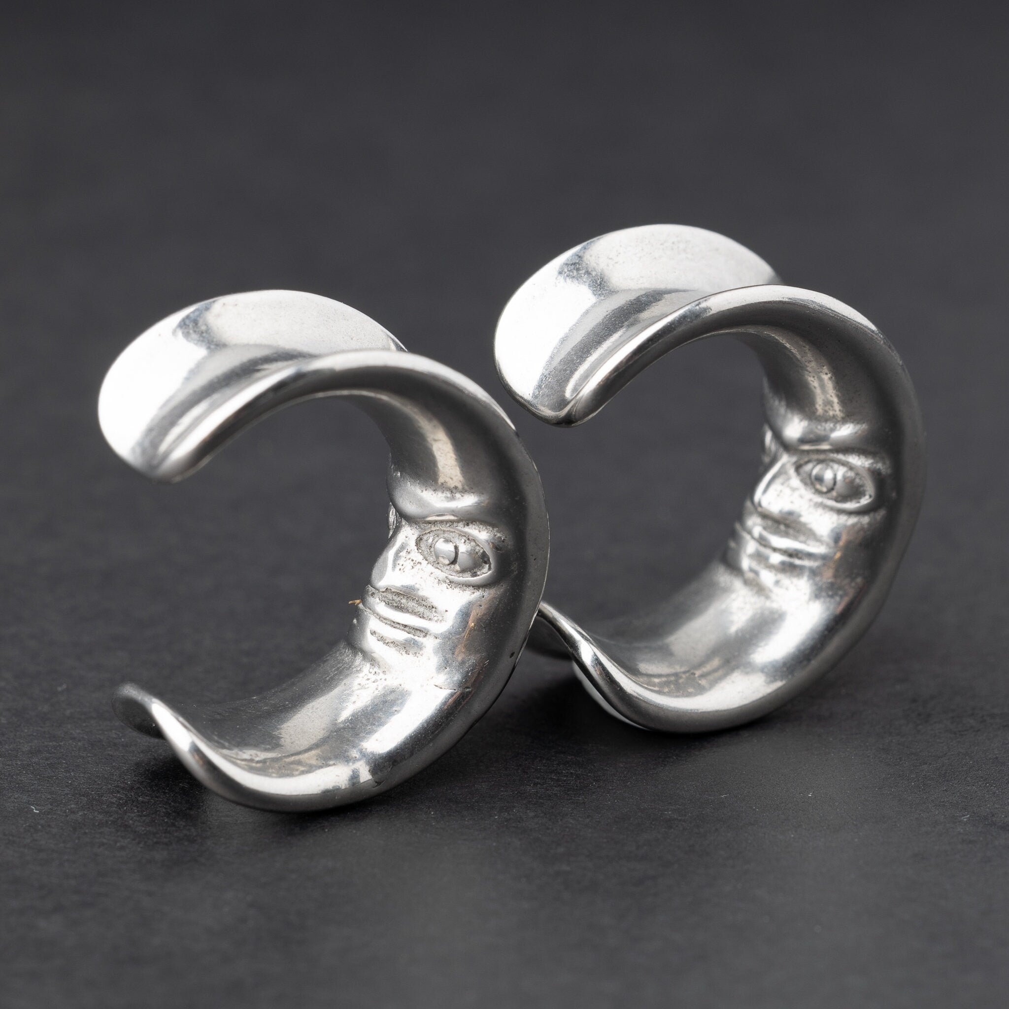 Close-up of a pair of silver crescent moon-shaped ear weights with a detailed face design, displayed on a dark background.