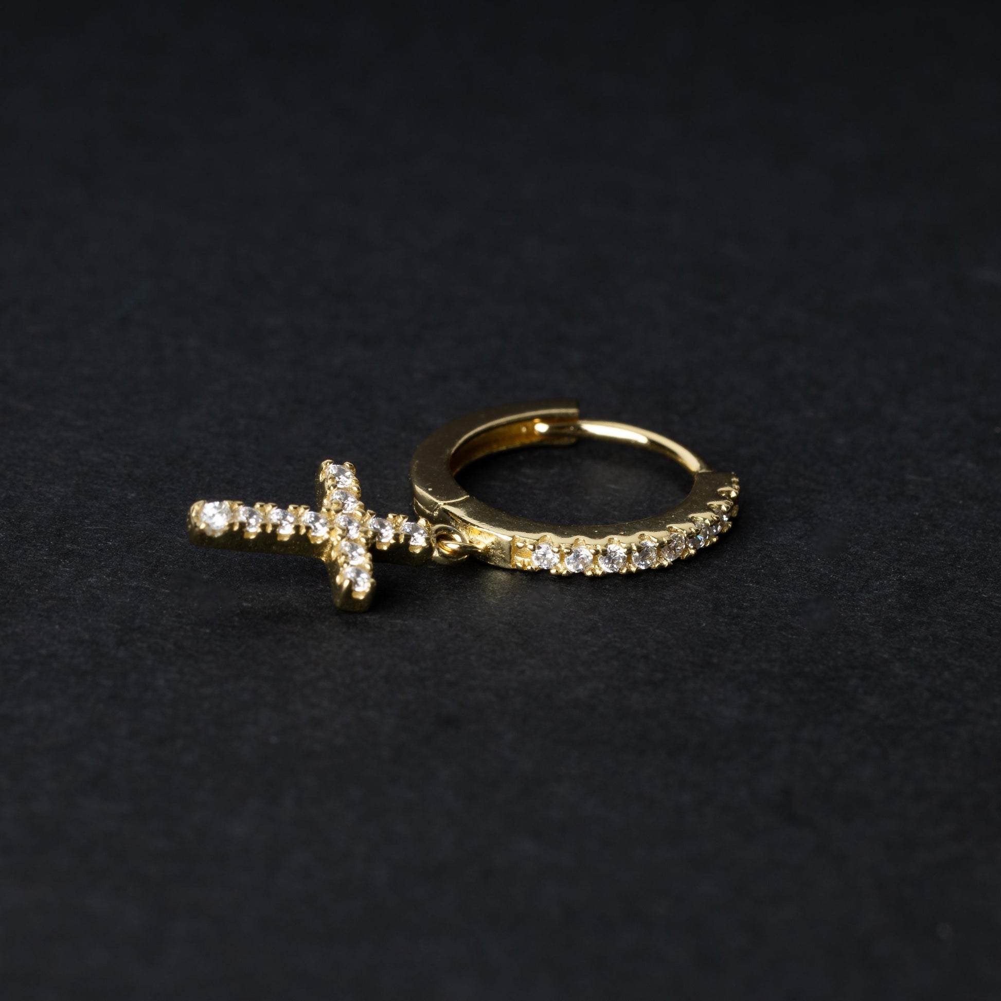 Gold hoop earring with cubic zirconia accents and a dangling cross charm, displayed on a dark background.