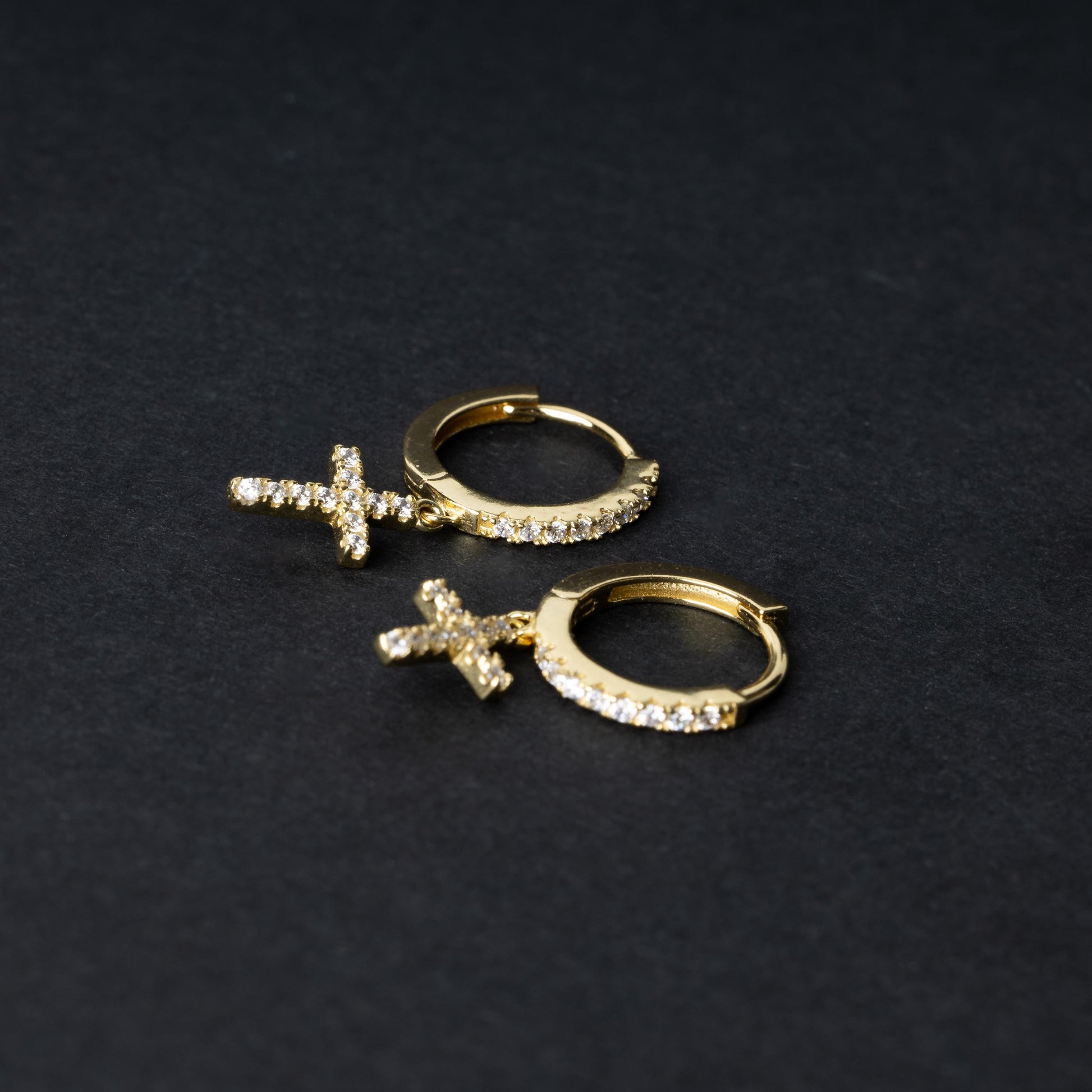 Gold hoop earrings with cubic zirconia accents and a dangling cross charm, displayed on a dark background.