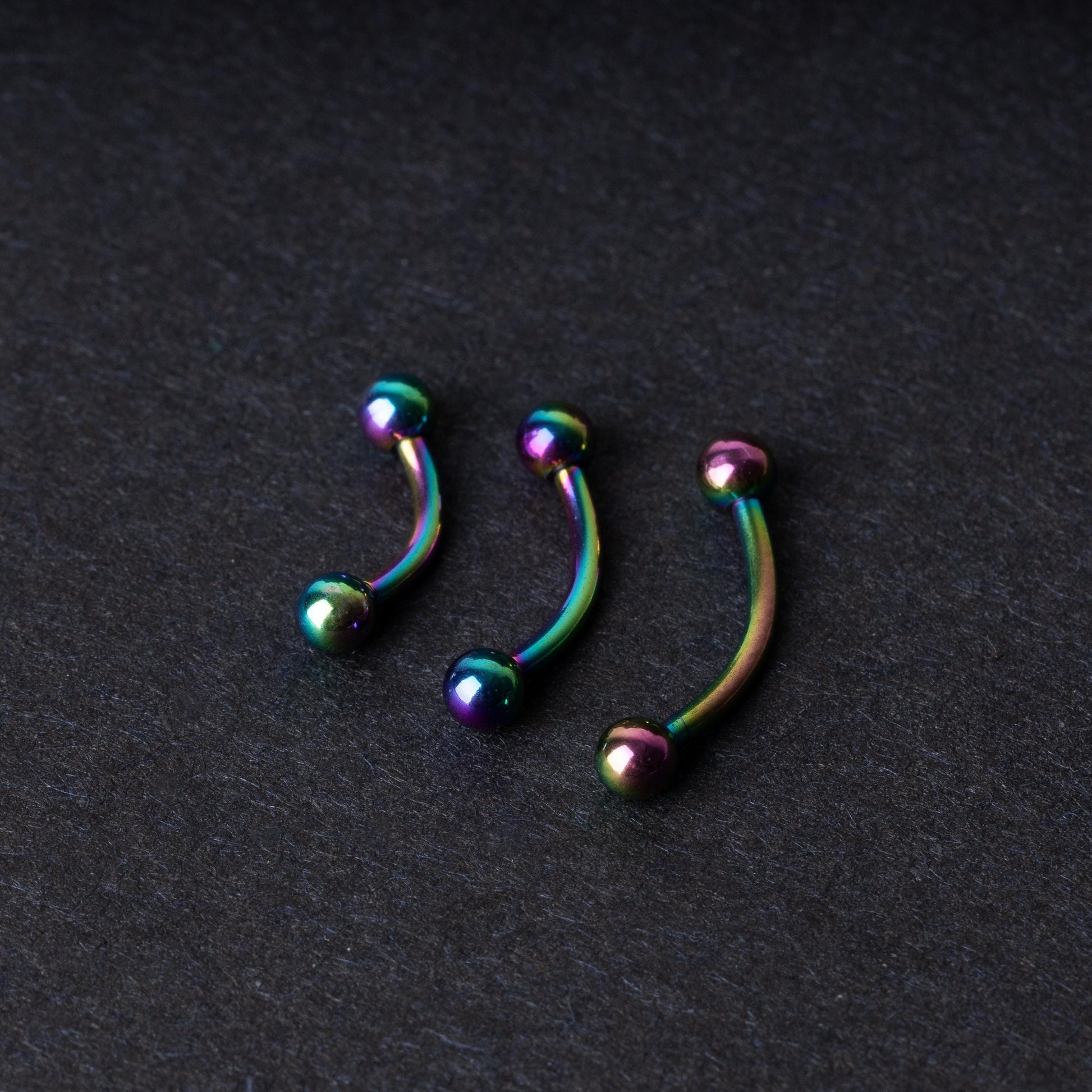 Three curved titanium barbells with a rainbow iridescent finish displayed on a dark textured background, highlighting their vivid colors and smooth finish.