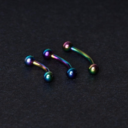 Three curved titanium barbells with a rainbow iridescent finish displayed on a dark textured background, highlighting their vivid colors and smooth finish.
