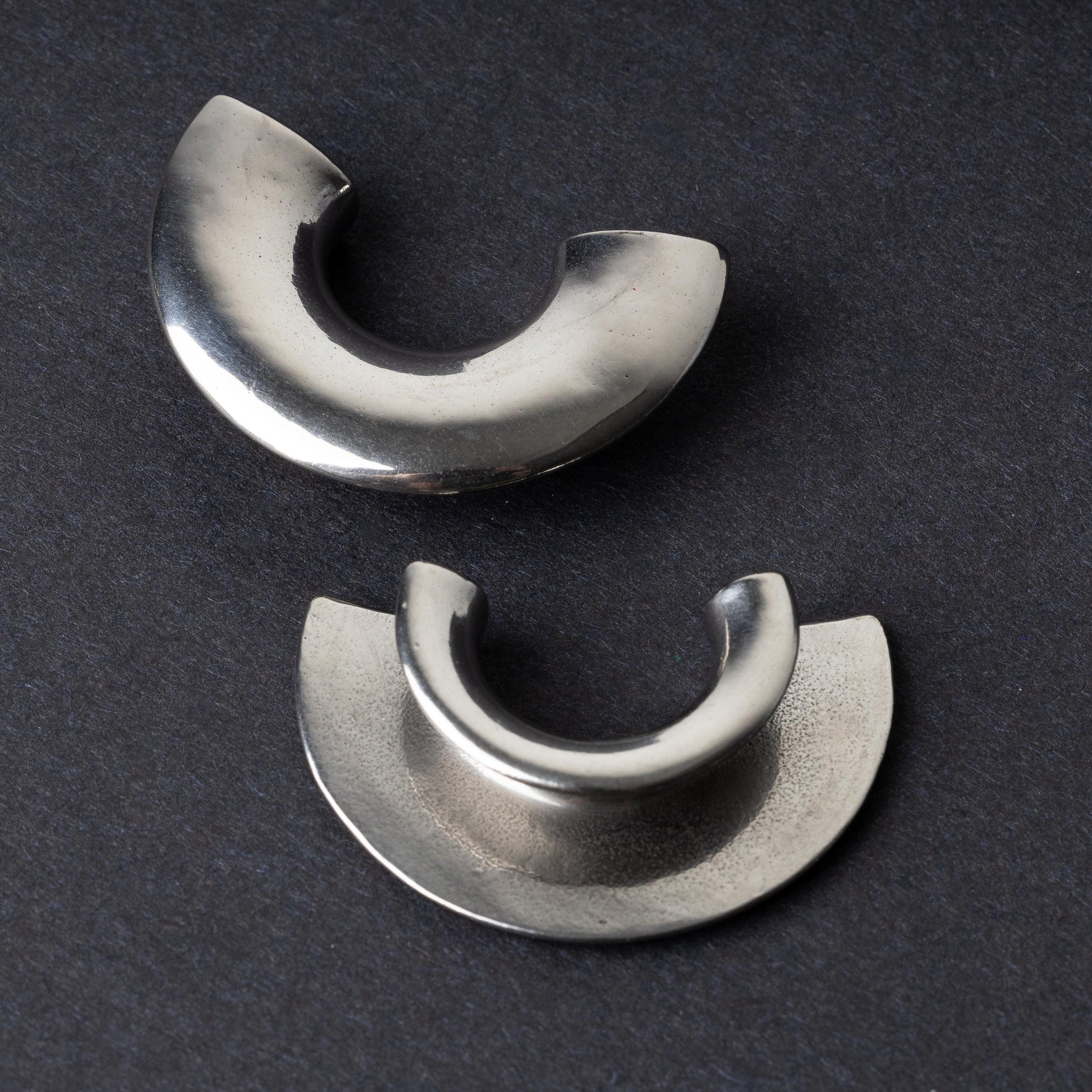 Close-up of two crescent-shaped stainless steel ear weights with a smooth, polished finish, displayed on a dark background.