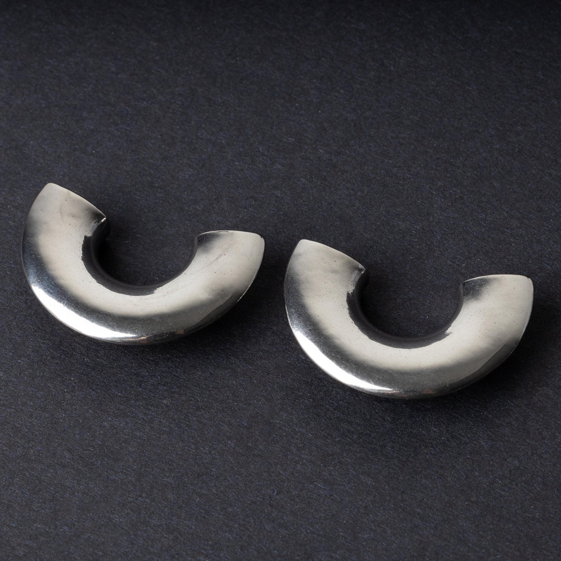 Close-up of two crescent-shaped stainless steel ear weights with a smooth, polished finish, displayed on a dark background.