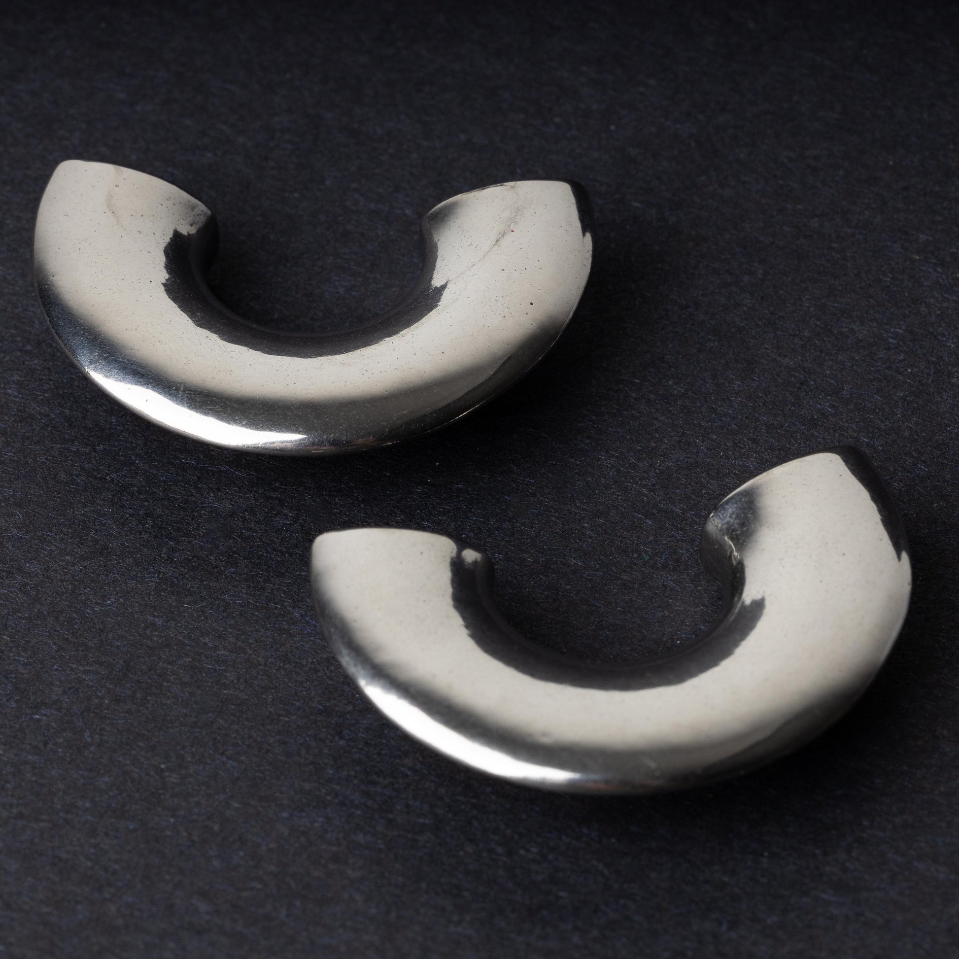 Close-up of two crescent-shaped stainless steel ear weights with a smooth, polished finish, displayed on a dark background.