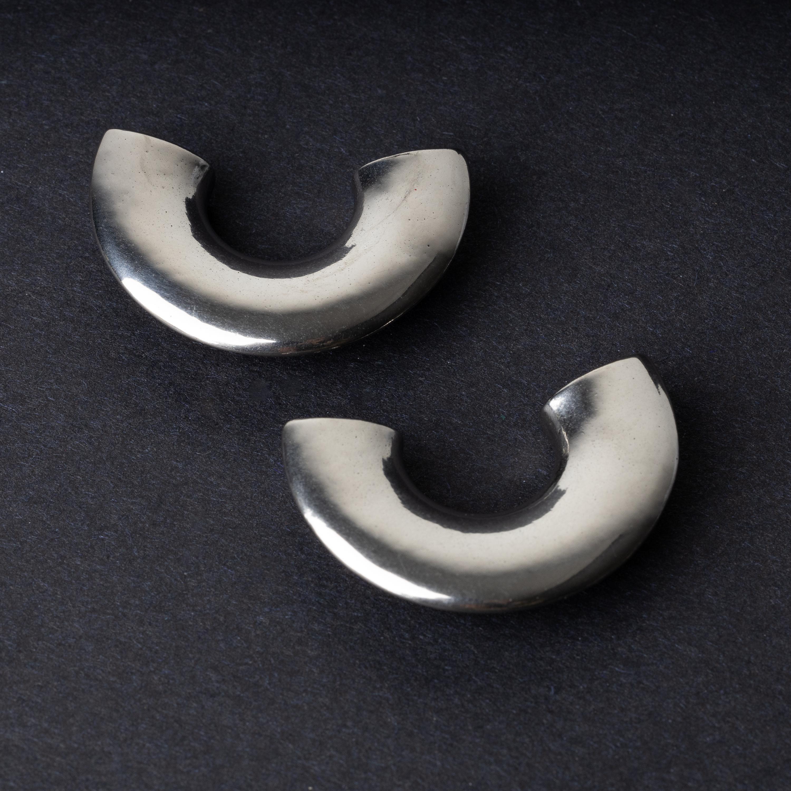 Close-up of two crescent-shaped stainless steel ear weights with a smooth, polished finish, displayed on a dark background.