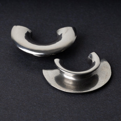 Close-up of two crescent-shaped stainless steel ear weights with a smooth, polished finish, displayed on a dark background.