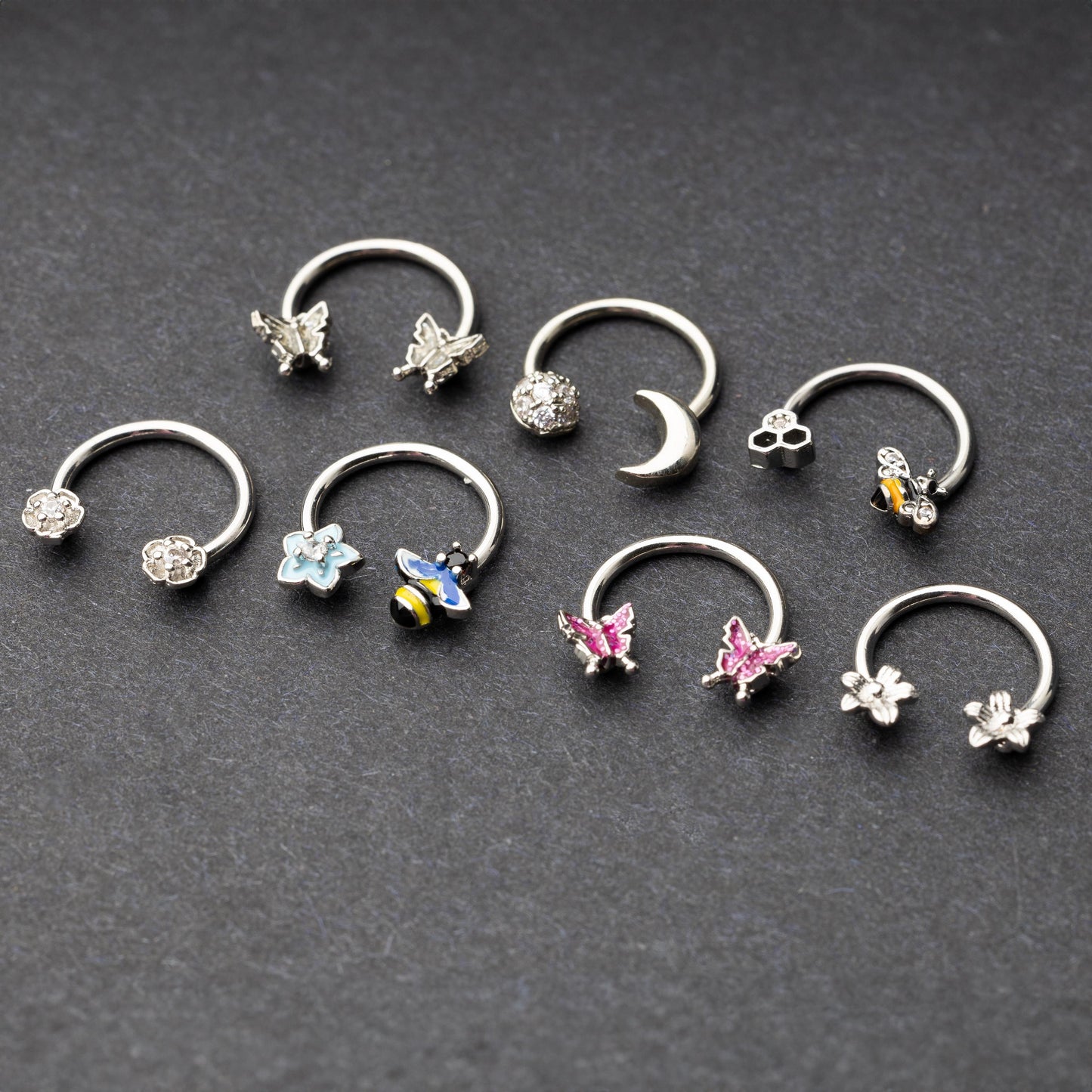 Silver horseshoe rings featuring various charms, including butterflies, bees, moons, and flowers, displayed on a dark background.