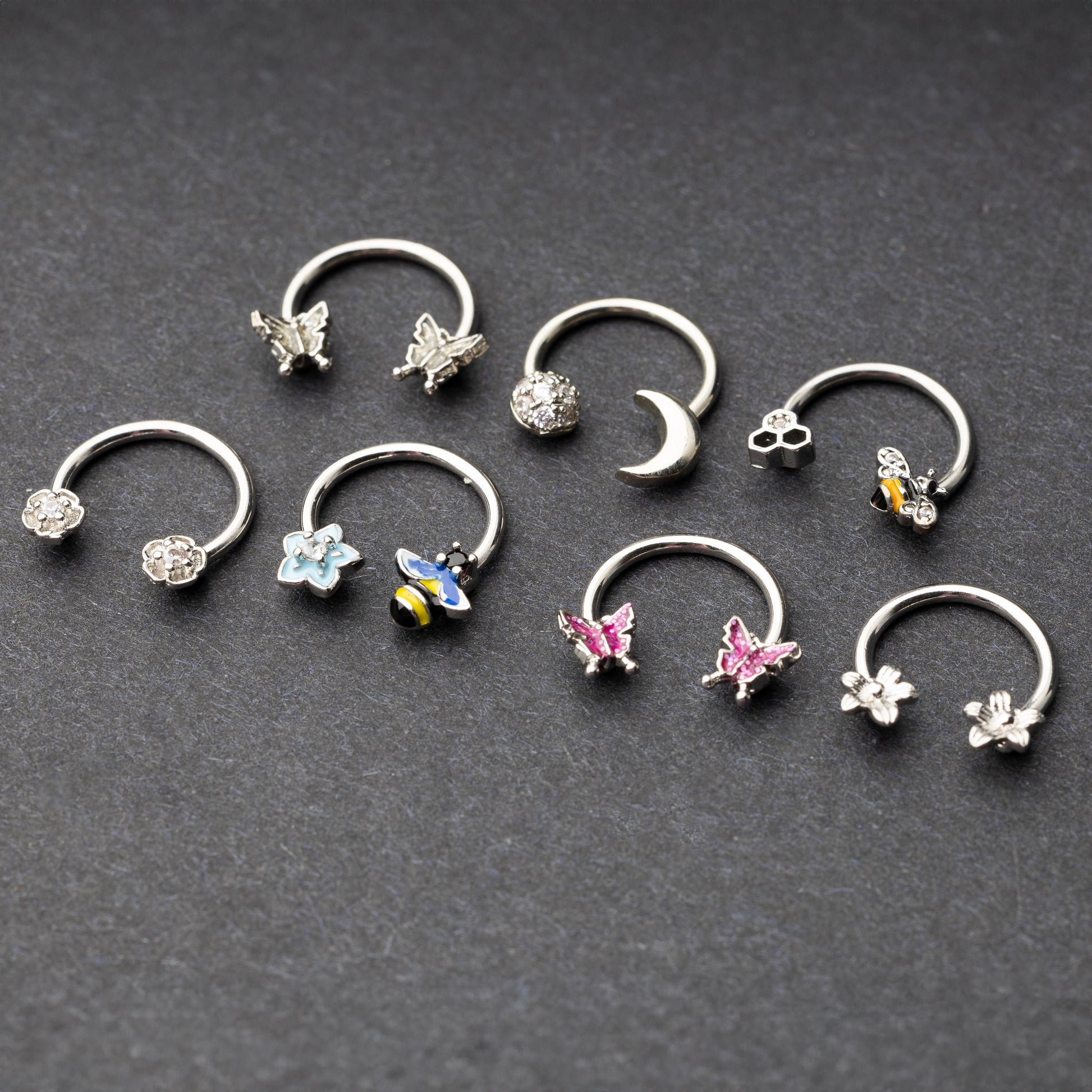 Silver horseshoe rings featuring various charms, including butterflies, bees, moons, and flowers, displayed on a dark background.